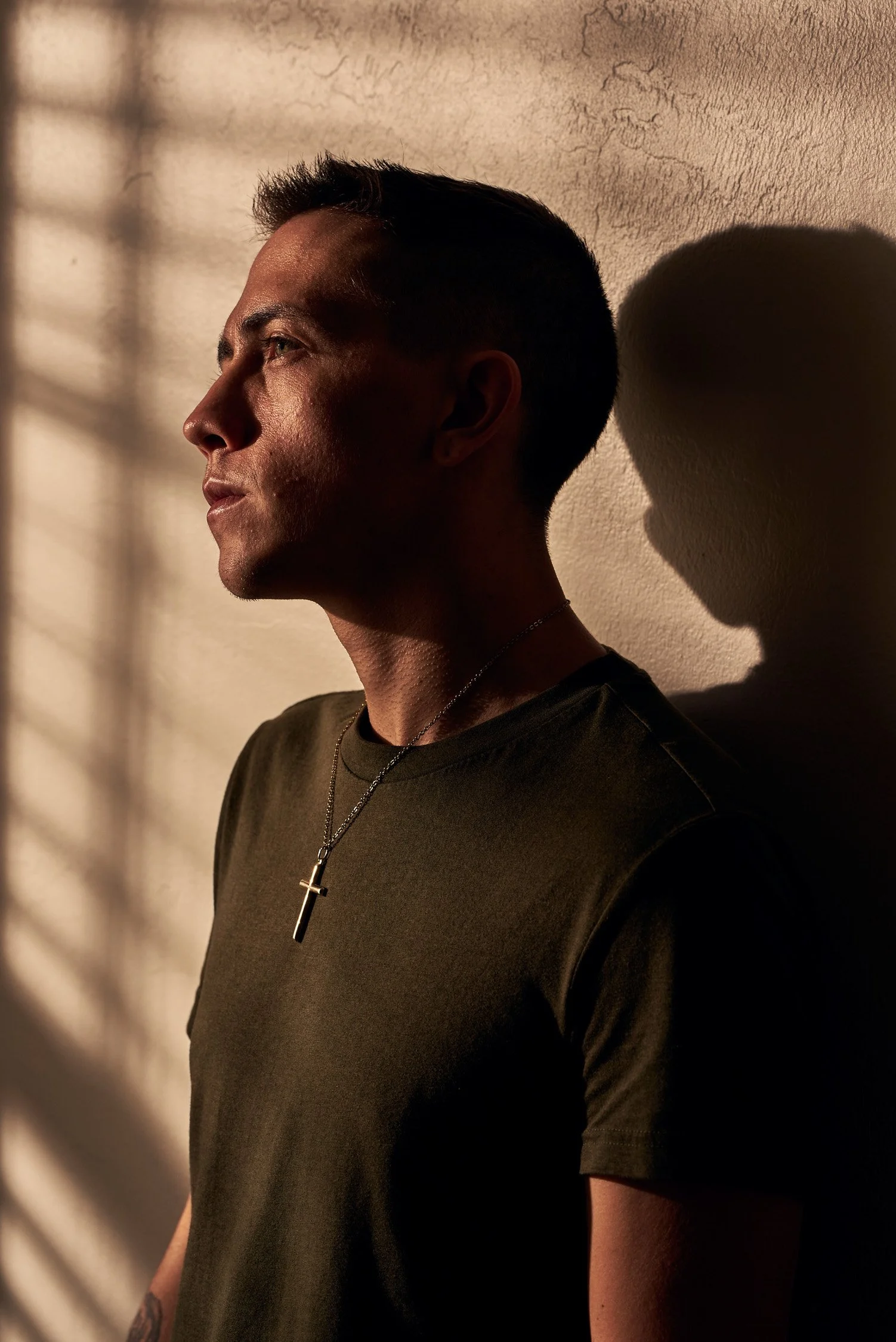 Side profile of a man with short dark hair, wearing a dark shirt and a silver cross necklace, standing against a textured wall with shadow patterns.