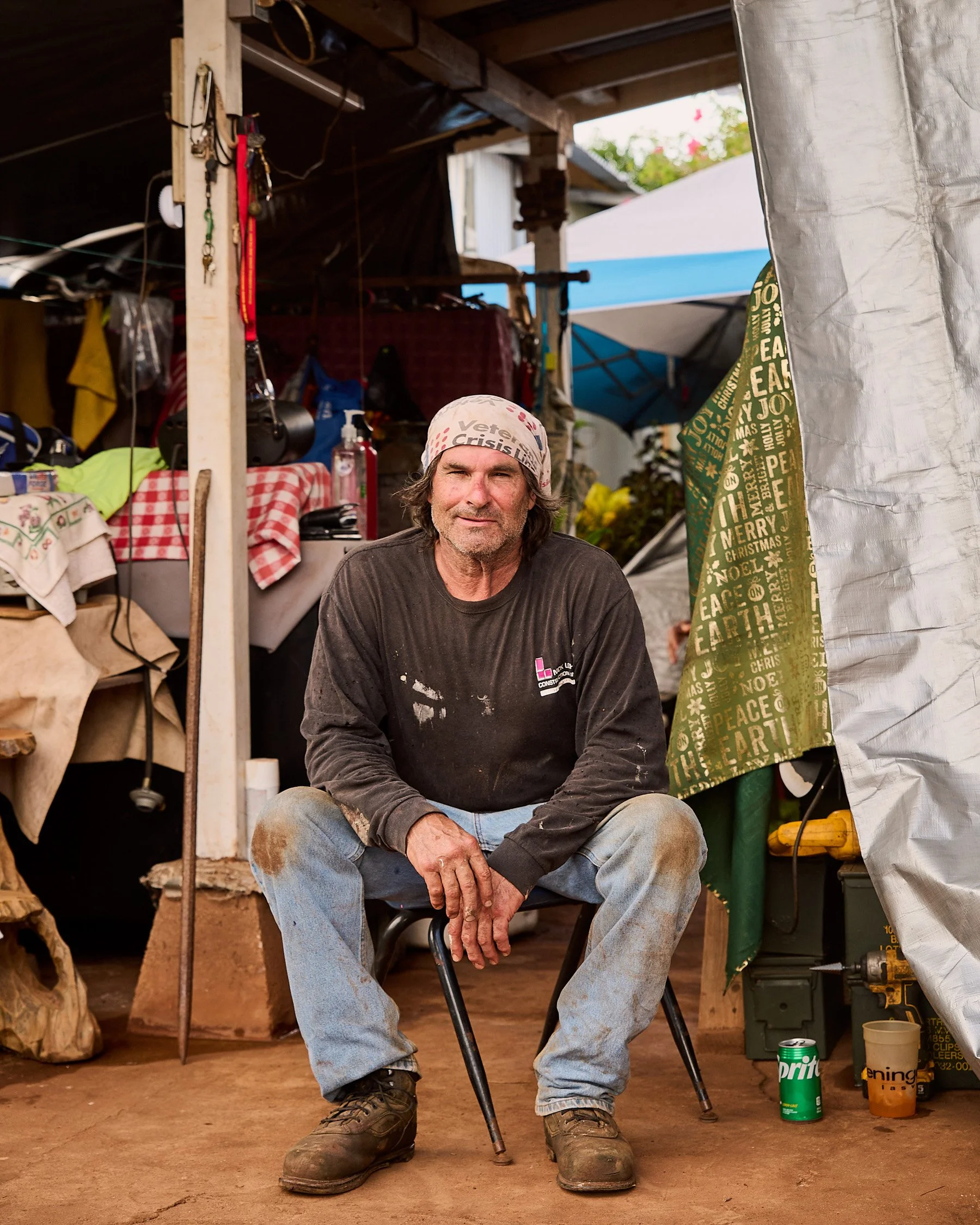 A man sitting on a chair inside a workshop or tent, with work stains on his clothing, wearing a bandana that reads 'Veterans Crisis Line'.