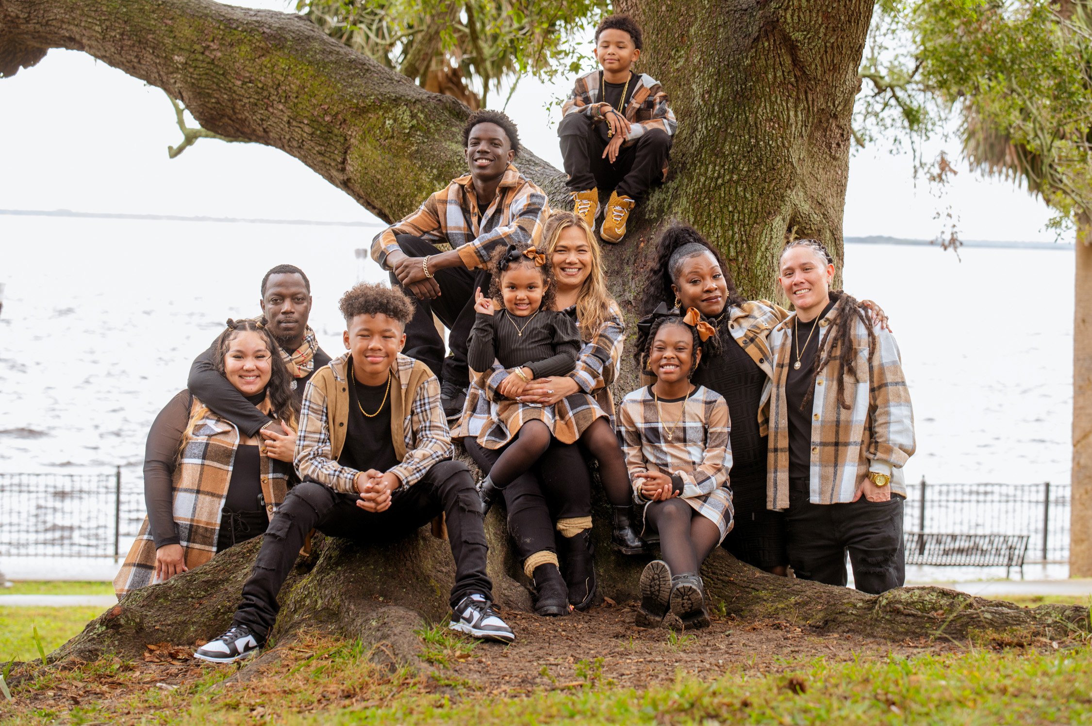 A group of diverse children and a woman sitting and standing around a large tree by a body of water, all wearing coordinated plaid outfits and smiling.