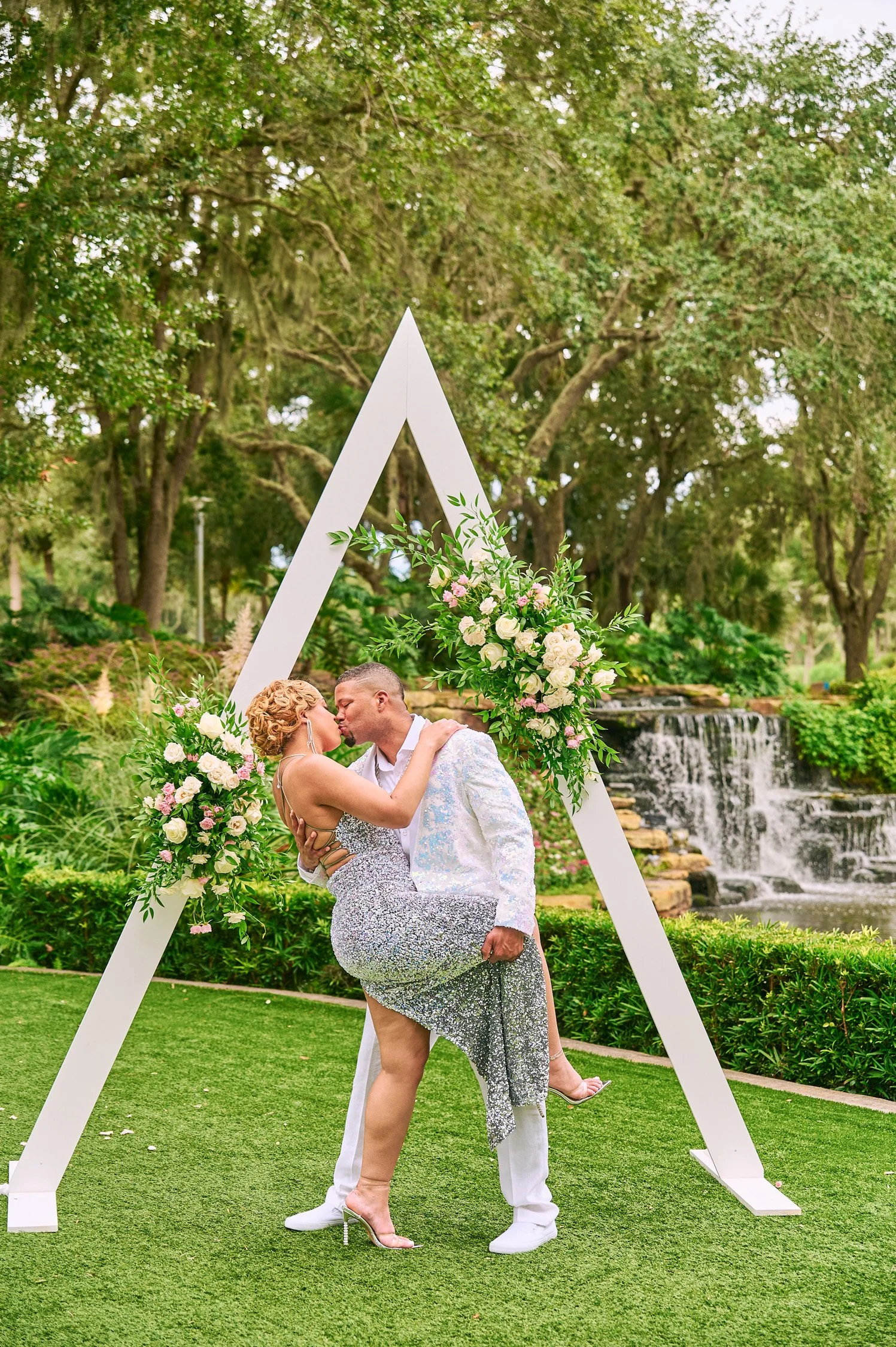 A couple kissing under a triangular wedding arch decorated with flowers outdoors, with greenery and a waterfall in the background.