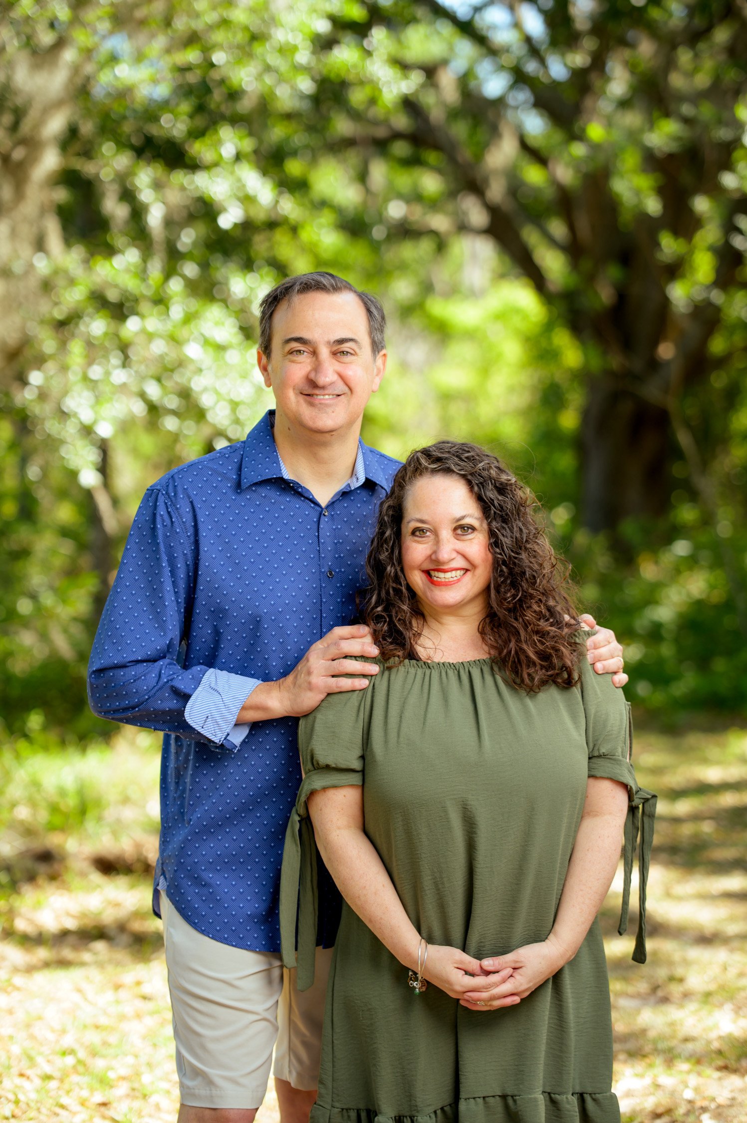 A man and woman standing outdoors in a wooded area, smiling at the camera. The man is wearing a blue long-sleeve shirt and khaki shorts, and the woman is wearing an olive green dress.