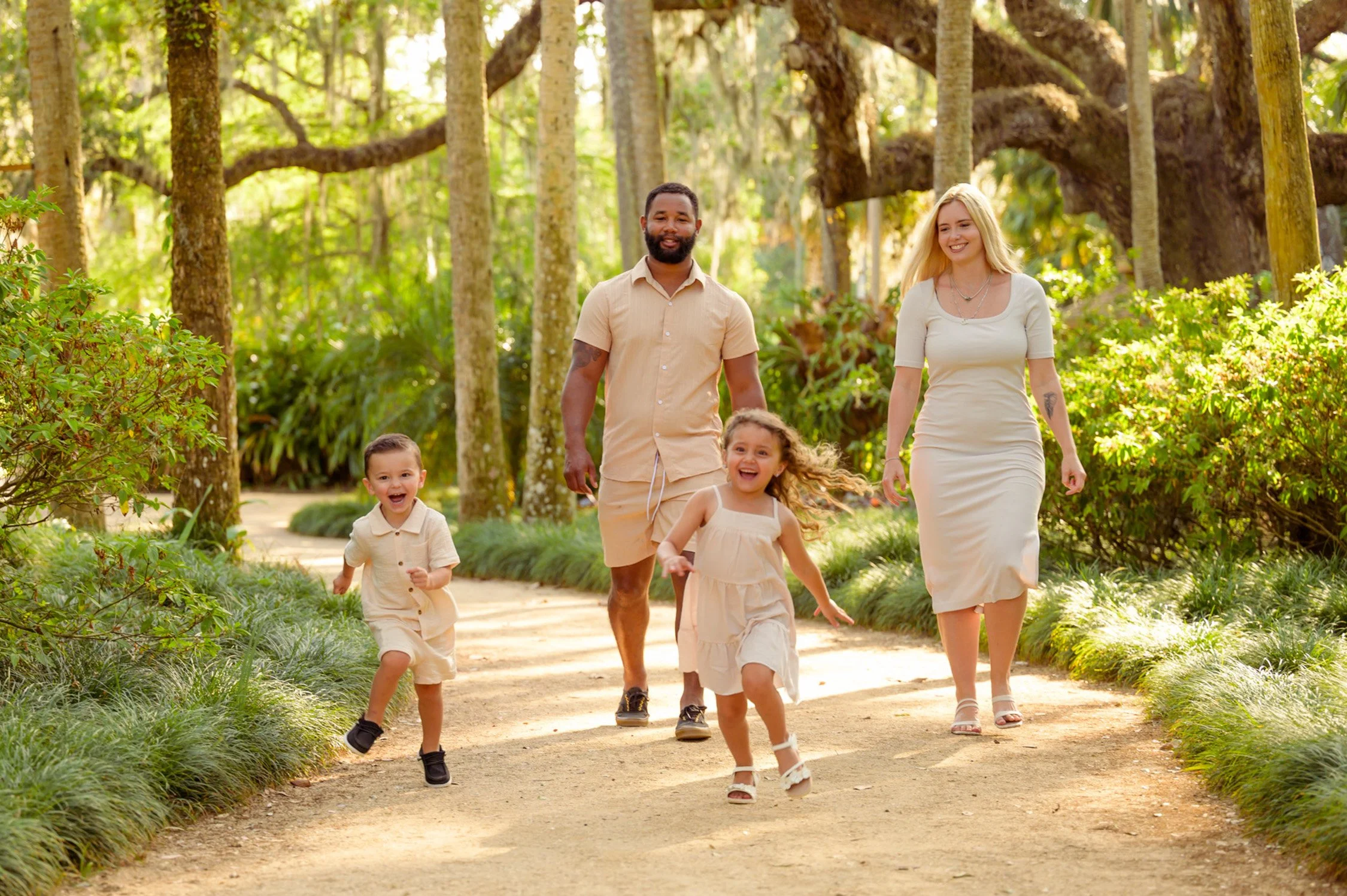 A family of four, two children and their parents, happily walking and running on a sunlit forest trail surrounded by greenery and trees.