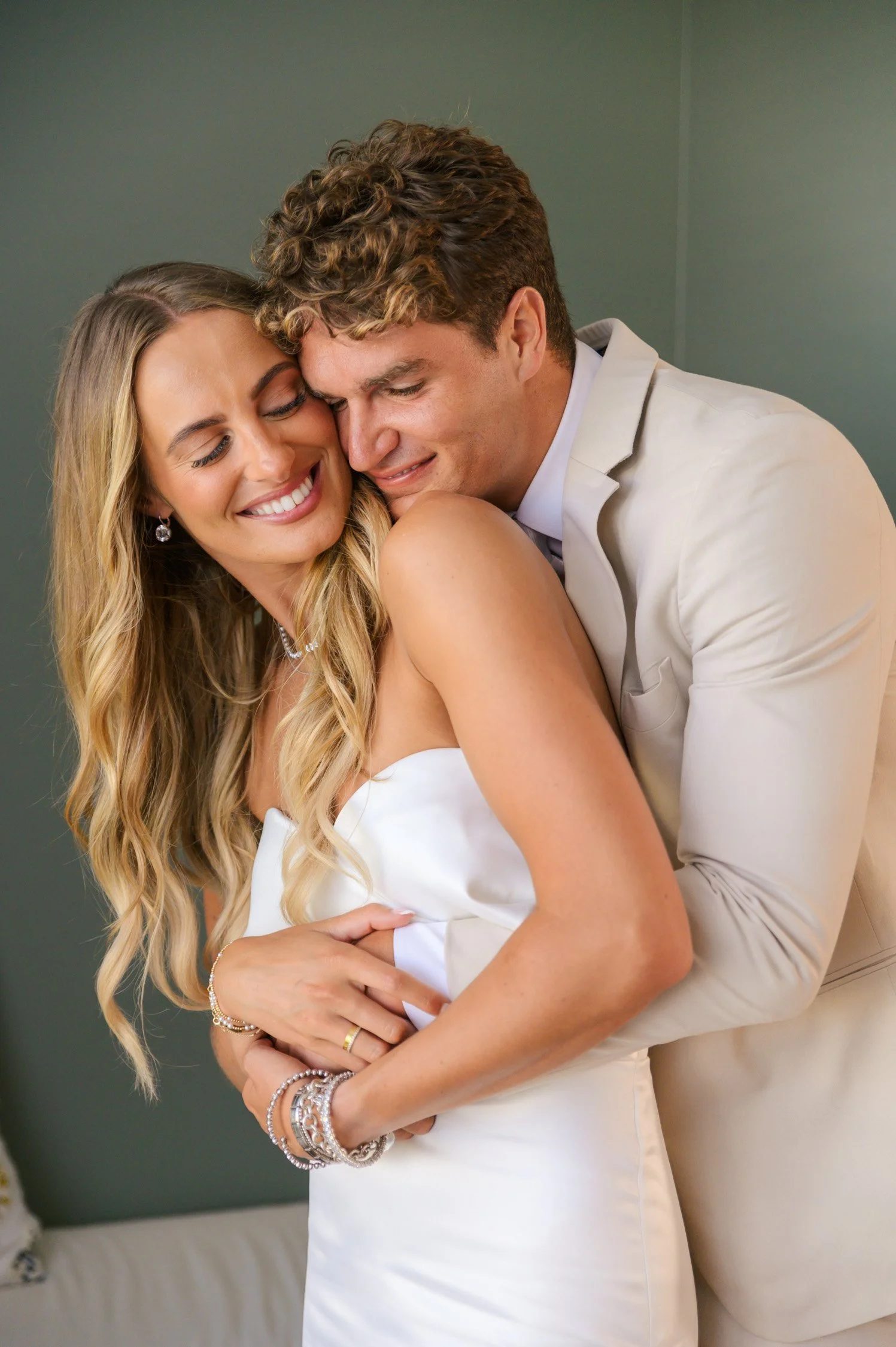A bride and groom hugging and smiling during their wedding, with the bride wearing a white wedding dress and the groom in a light-colored suit.