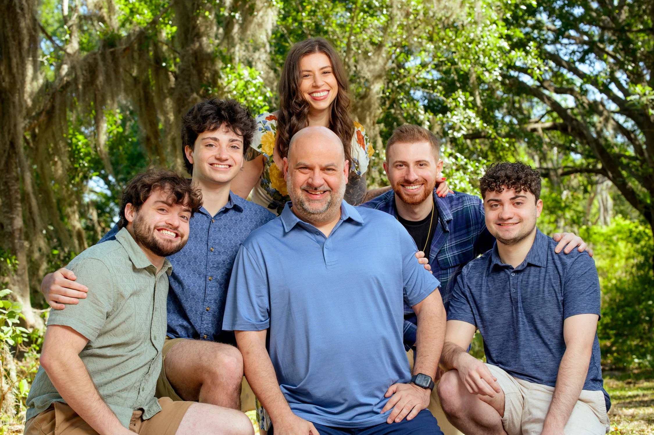 Group of seven people, five men and two women, smiling outdoors in a forested area, with green leaves and trees in the background.