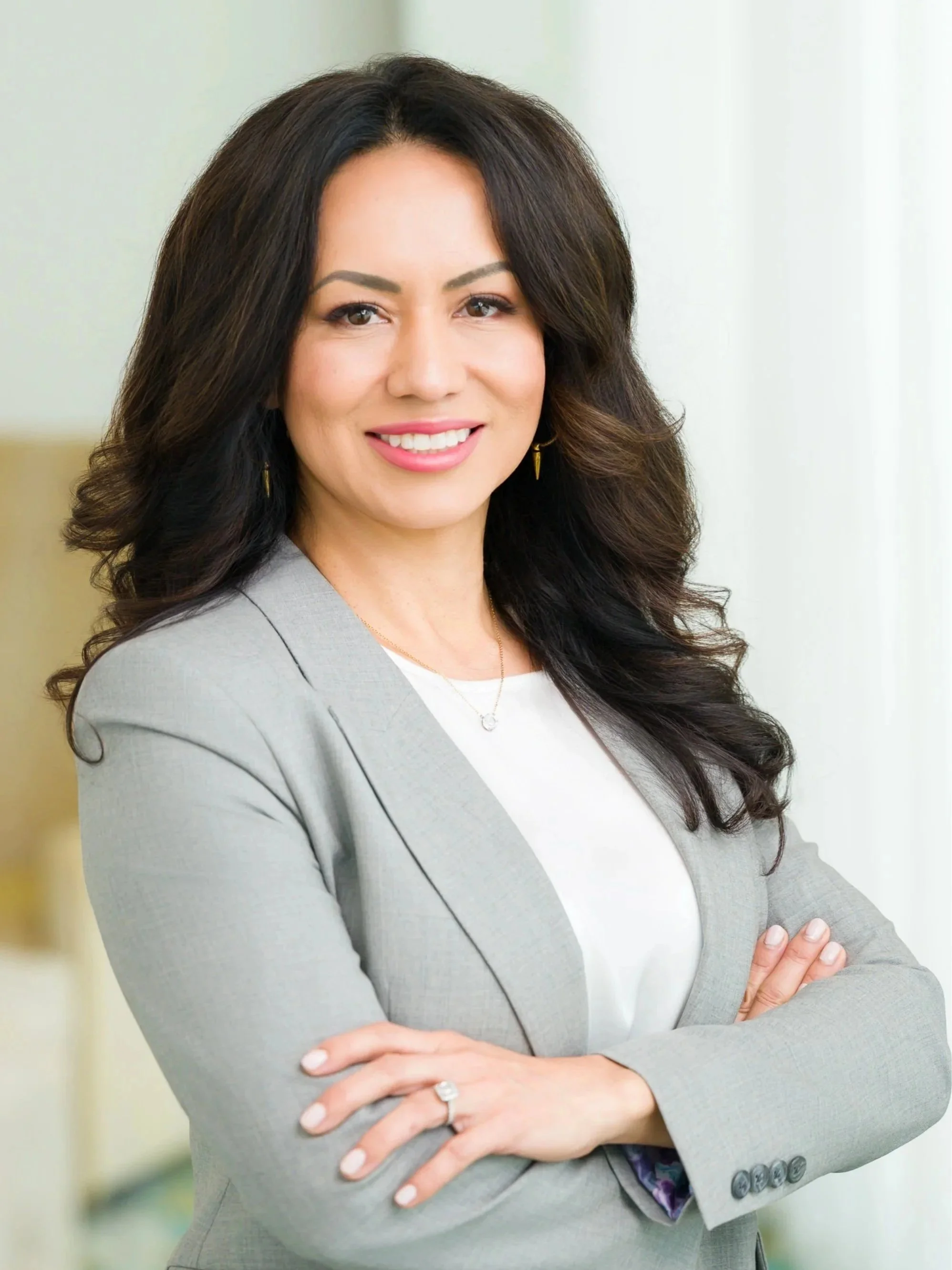 A professional woman with long dark hair, wearing a light gray blazer and white top, smiling with arms crossed, standing in a bright office.