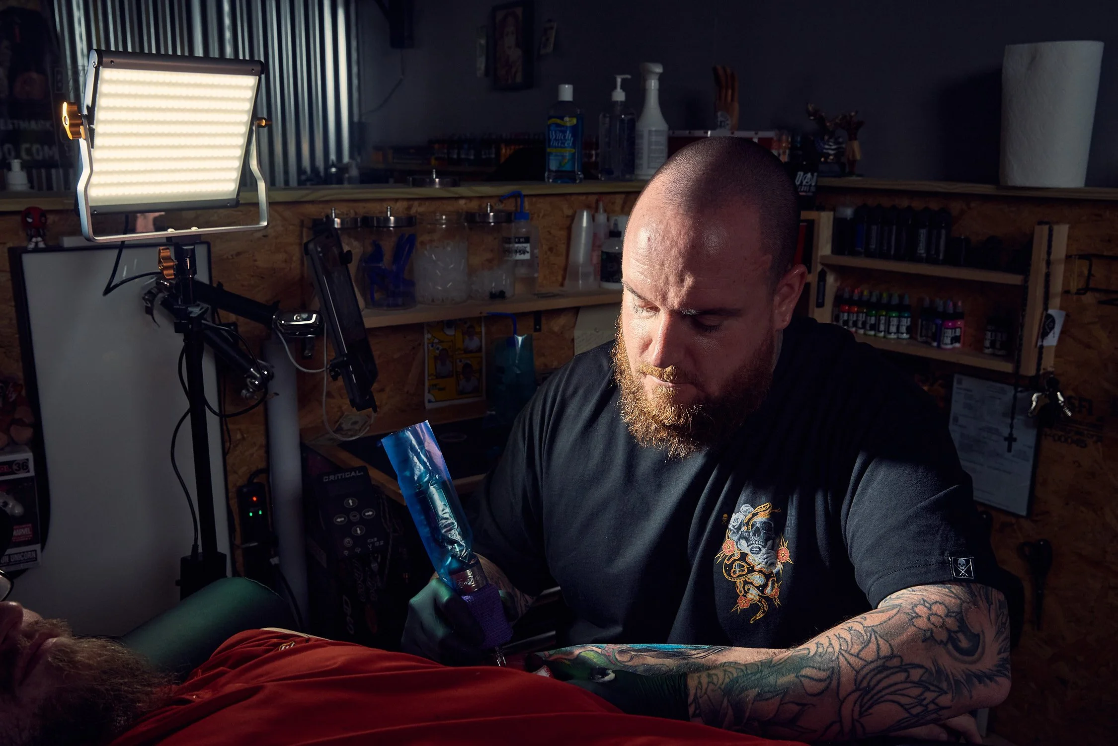 Tattoo artist working on a client's arm in a tattoo studio, with tattoo supplies and tools on shelves in the background.