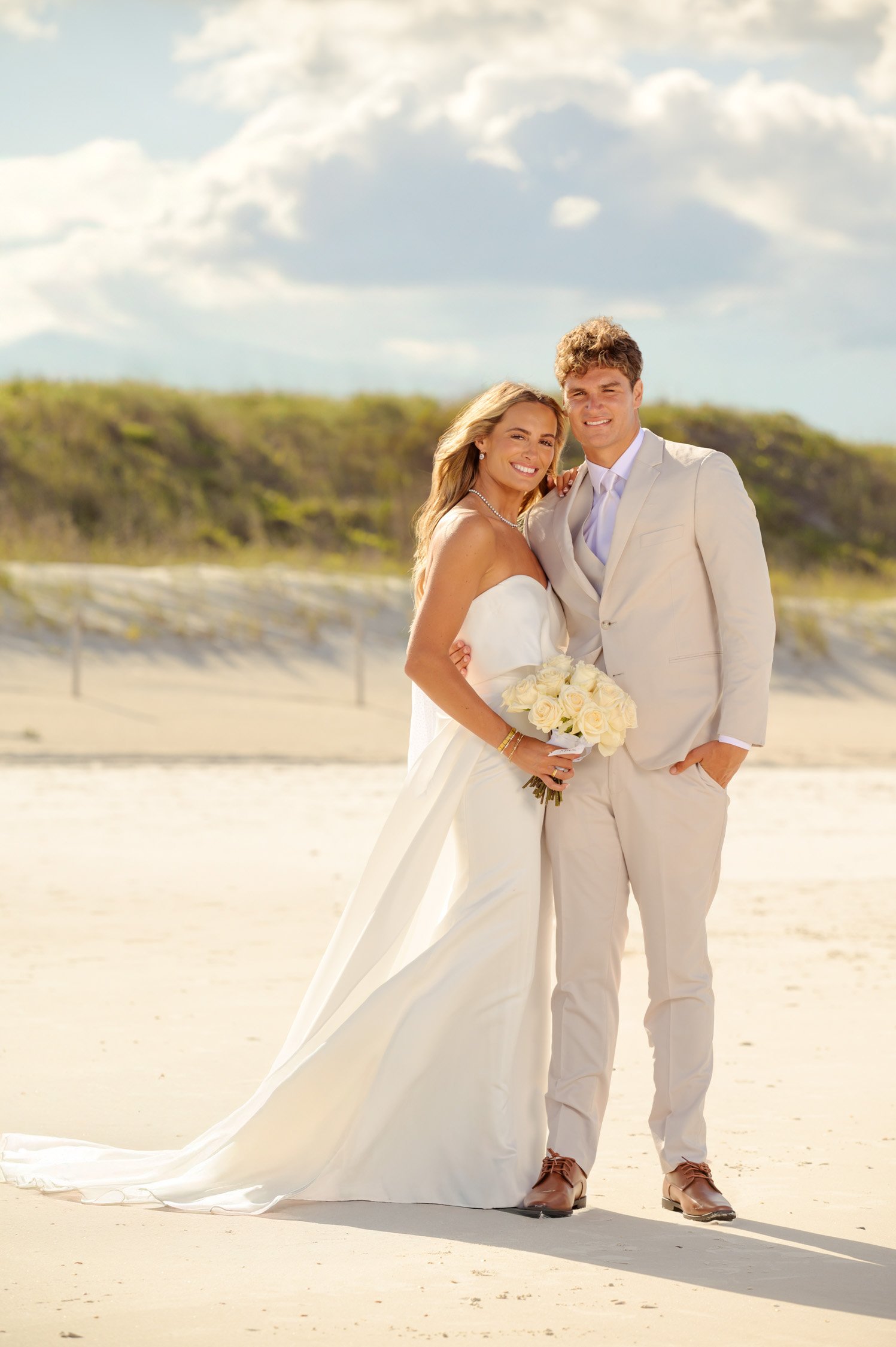A bride and groom standing on a beach, smiling, with the bride holding a bouquet of white roses.