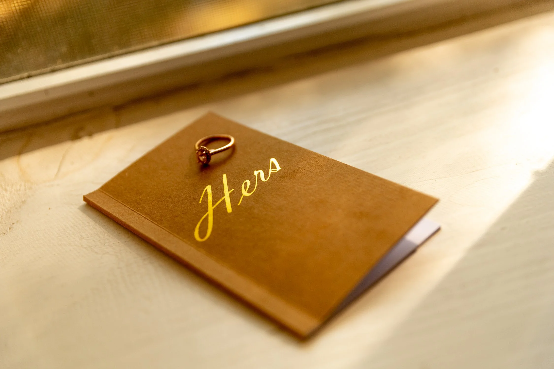 A rose gold engagement ring with a central diamond, placed on a brown card with the handwritten word 'Hers' in gold lettering, on a wooden surface.