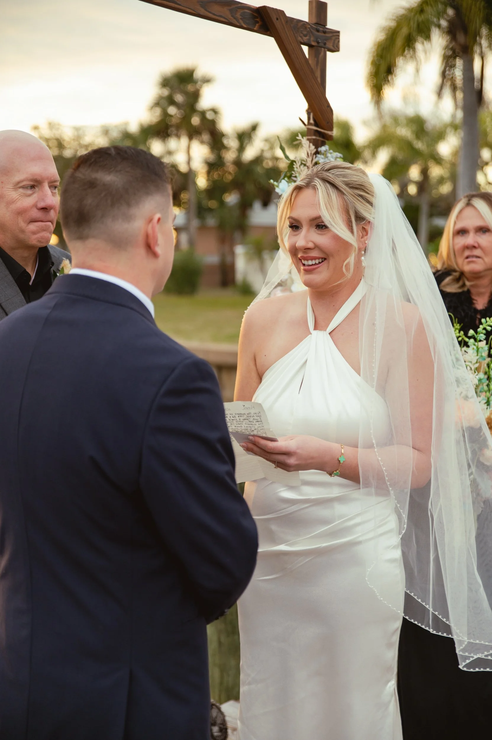A woman in a white wedding dress holding a note, smiling at a man in a suit during an outdoor wedding ceremony at sunset, with guests and palm trees in the background.