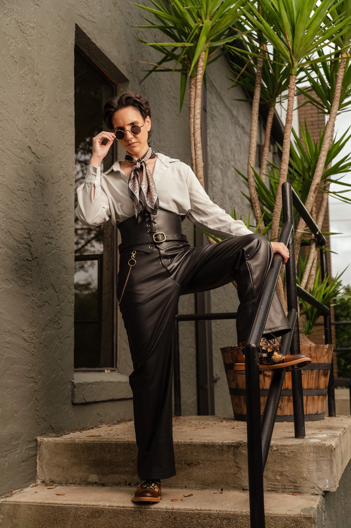 A woman dressed in a white shirt, black leather pants, and brown boots, wearing sunglasses and a scarf, stands on outdoor stairs with a railing. She is holding the railing with one hand and adjusting her sunglasses with the other. Behind her are tall