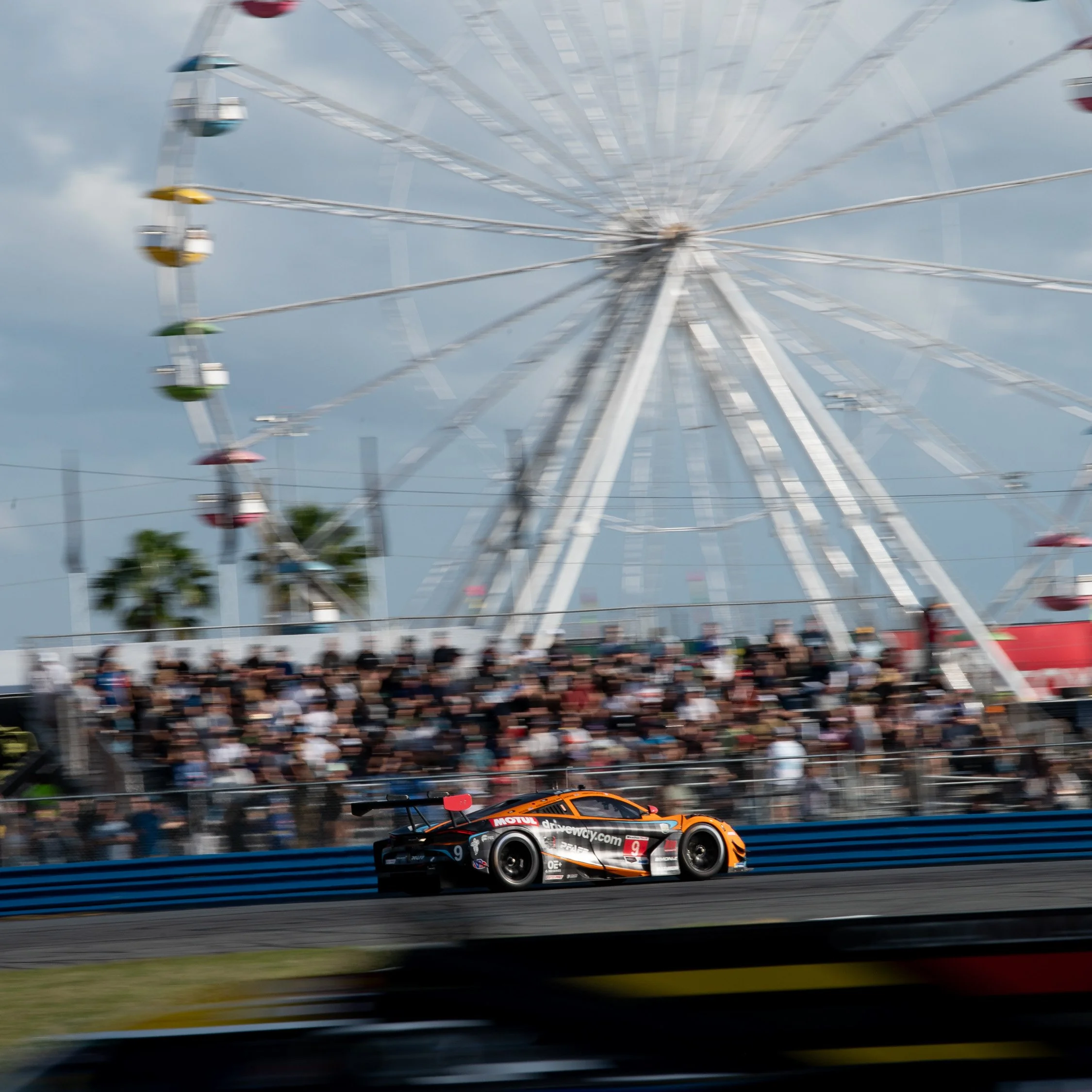 A race car on a track with a ferris wheel and spectators in the background.