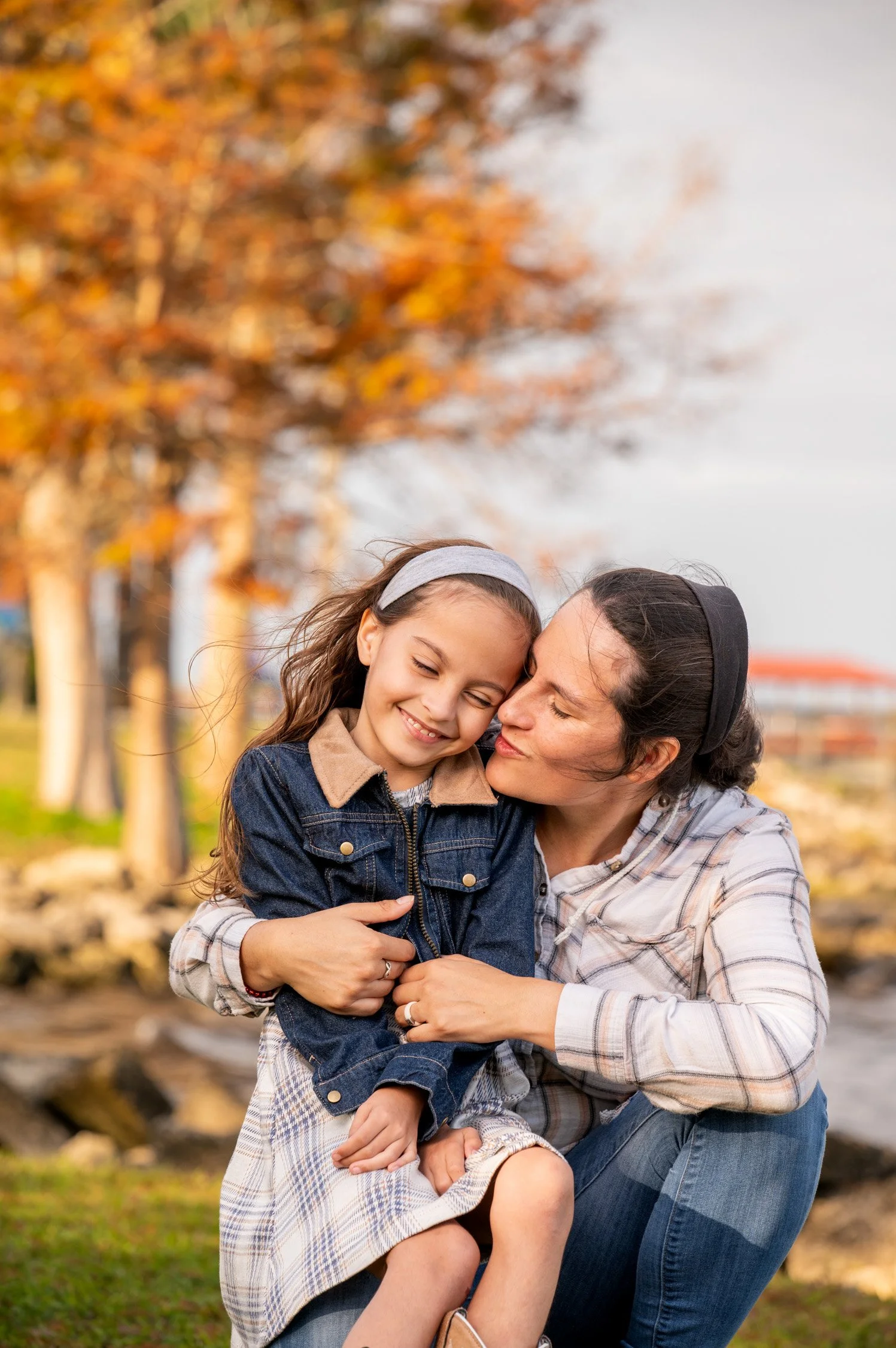 A woman is hugging a young girl outdoors near a body of water with autumn trees in the background, both smiling and enjoying each other's company.