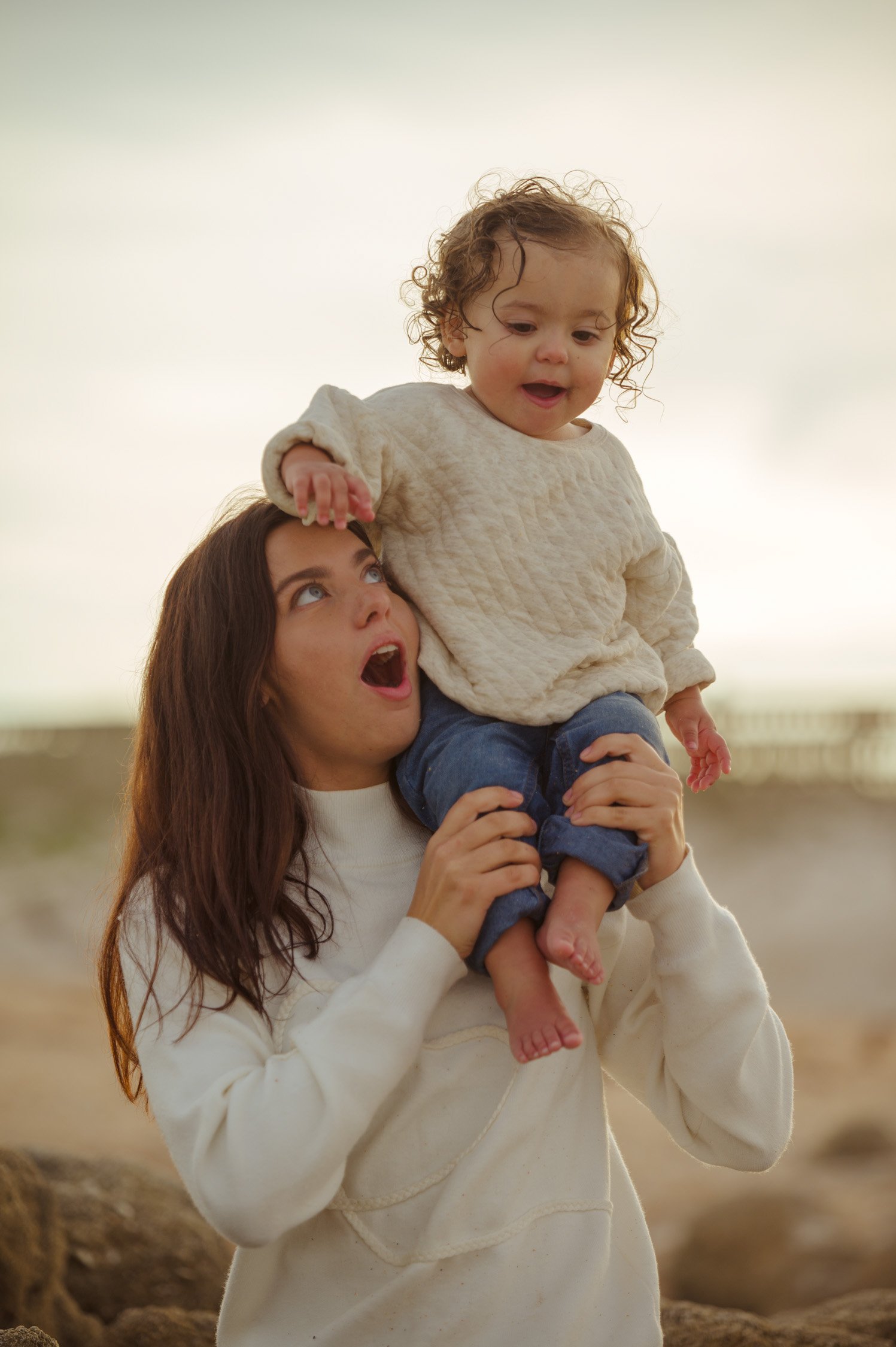 A woman with long brown hair holding a young girl with curly hair on her shoulders at the beach.