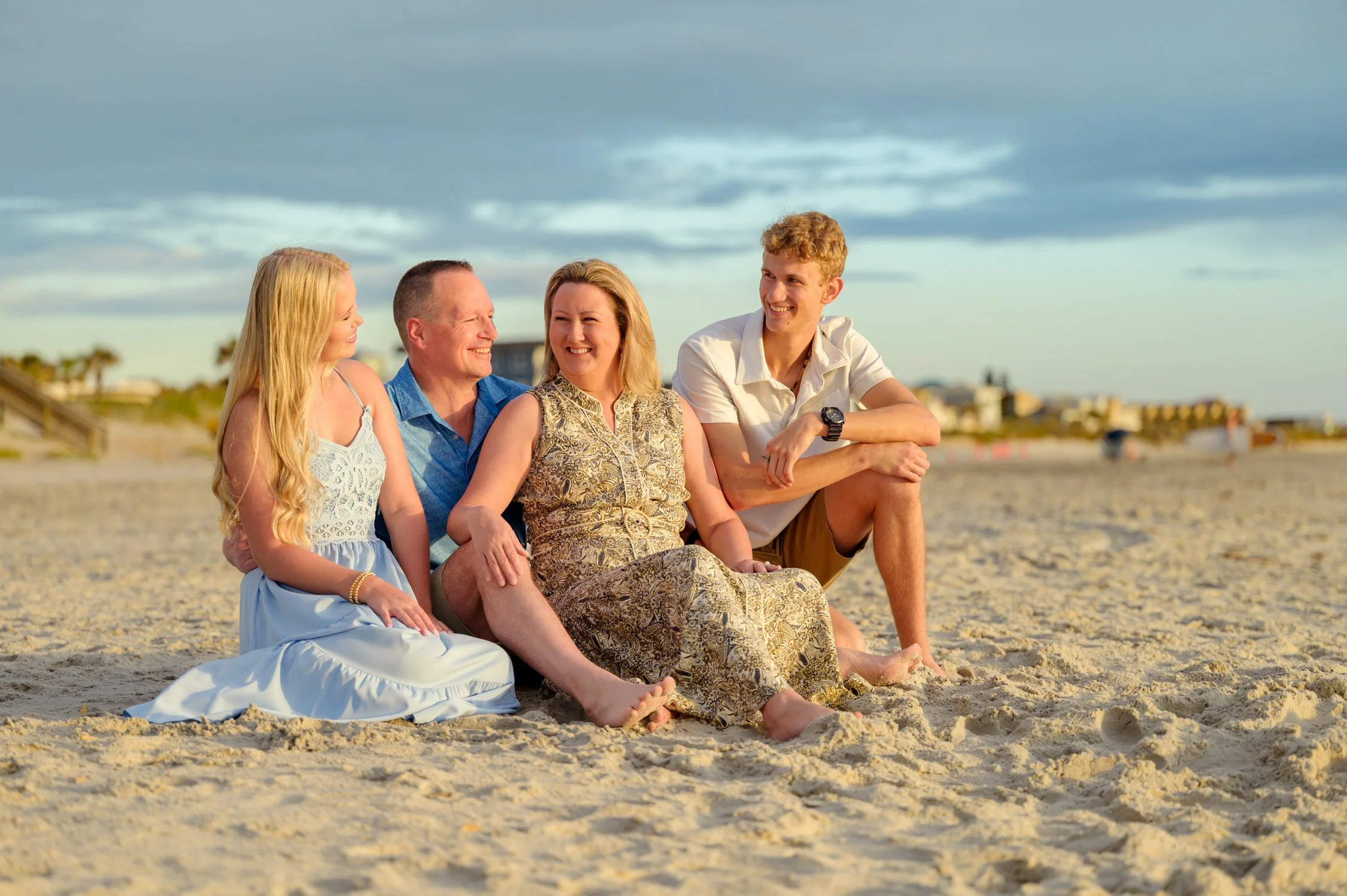 A family of five sitting on the sand at the beach, smiling and enjoying each other's company during sunset, with a cloudy sky and distant buildings in the background.