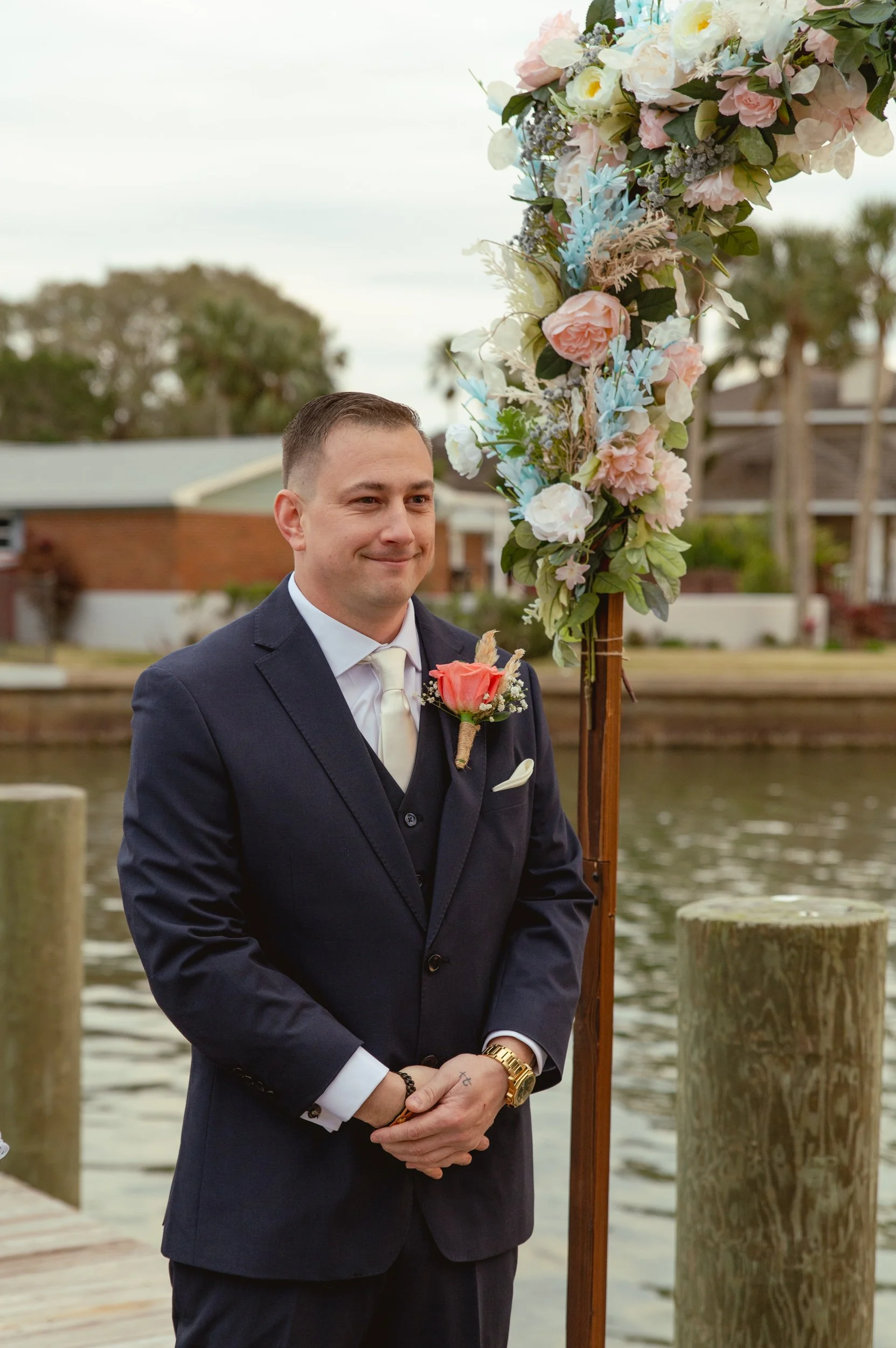 A groom in a navy suit standing by a floral wedding arch near a body of water.