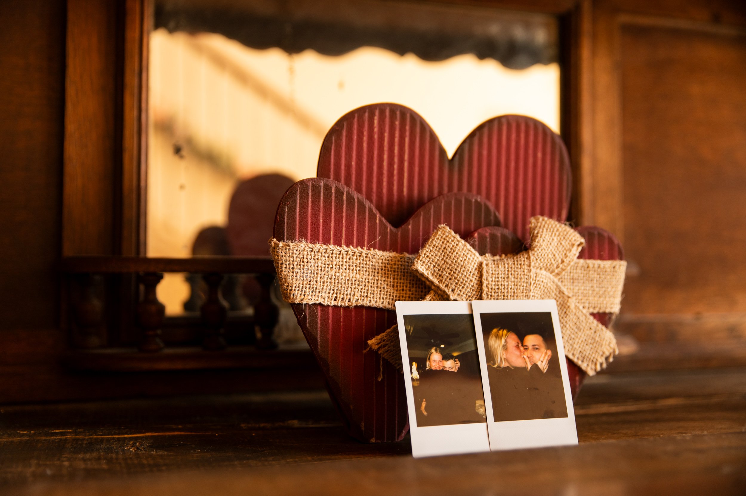 A decorated wooden table features a heart-shaped gift box wrapped with burlap ribbon and two instant photographs in front of it, with a mirror and wooden furniture in the background.