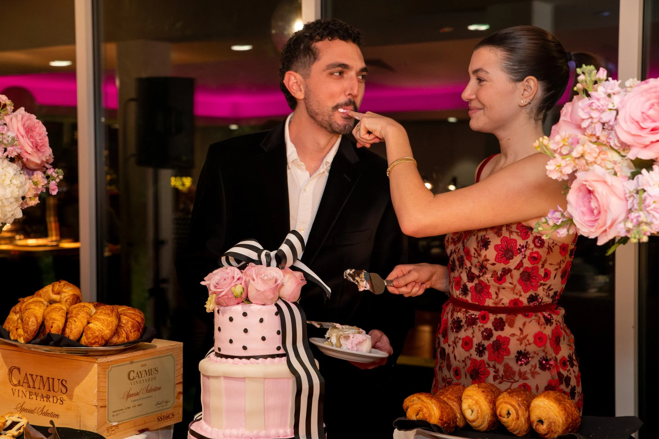 A woman feeds a man a piece of cake at a celebration party, with a decorated cake, pastries, and flowers on the table.