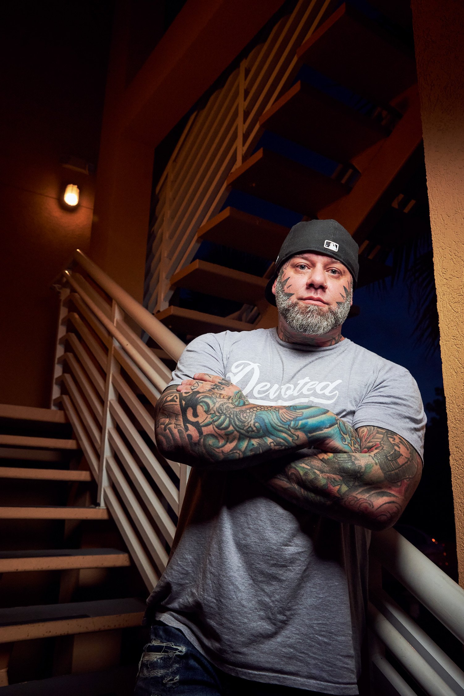 A tattooed man with a beard, wearing a gray T-shirt and a black cap, stands with arms crossed in front of a staircase at night.