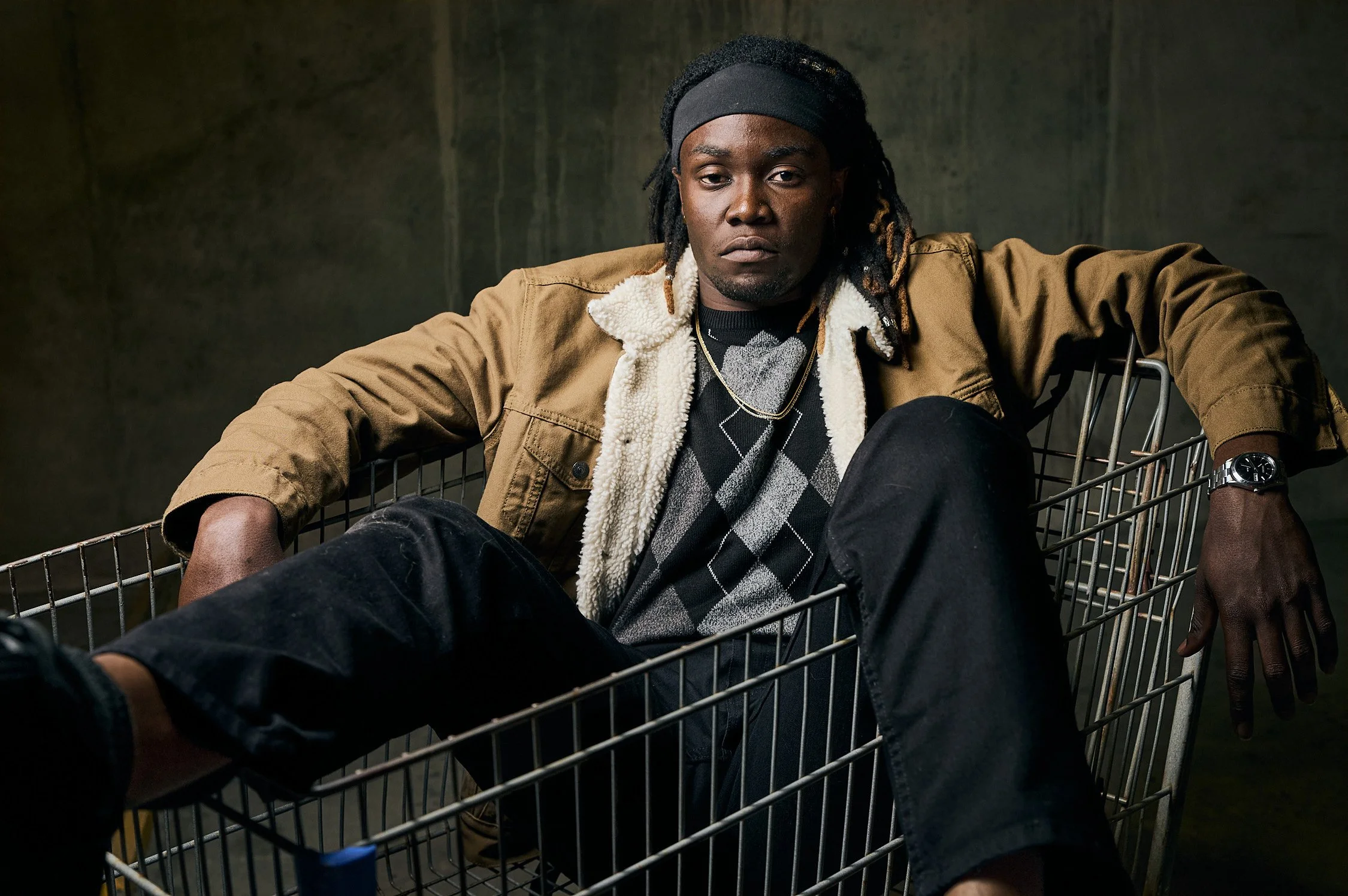 A man sitting in a shopping cart with a serious expression, wearing a brown jacket, argyle sweater, black pants, a watch, and a headband, against a dark background.