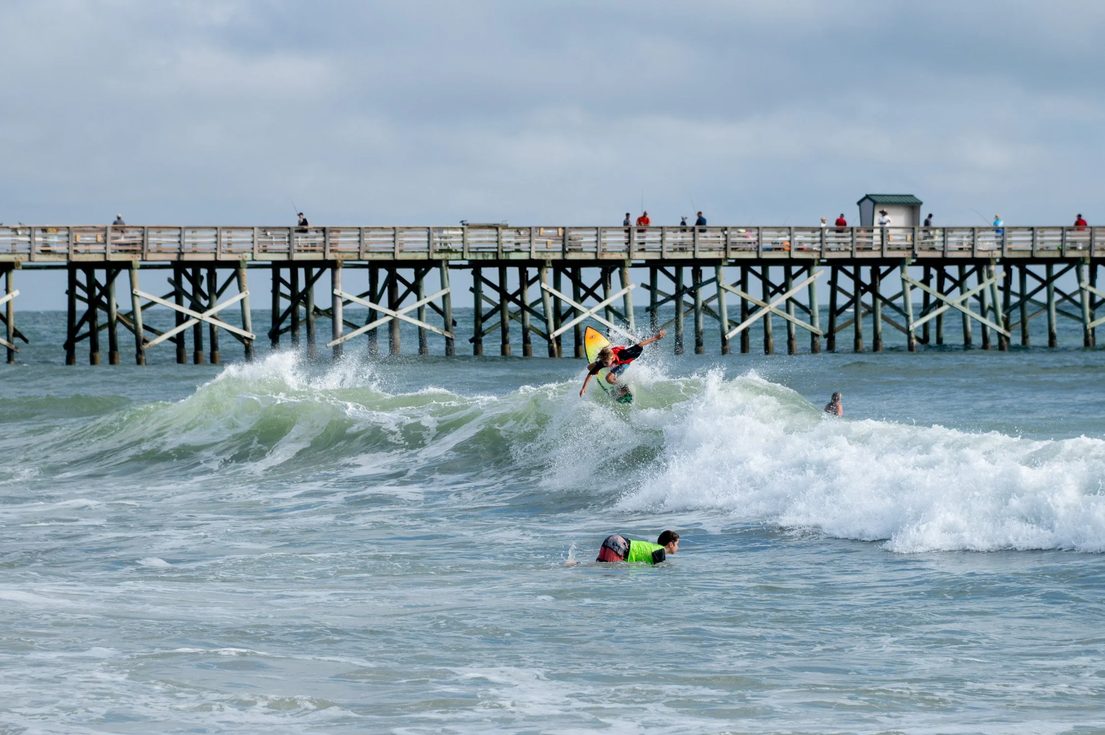 A surfer in a red and black wetsuit riding a wave near a pier with several people fishing and walking on it, with two swimmers in the water below.