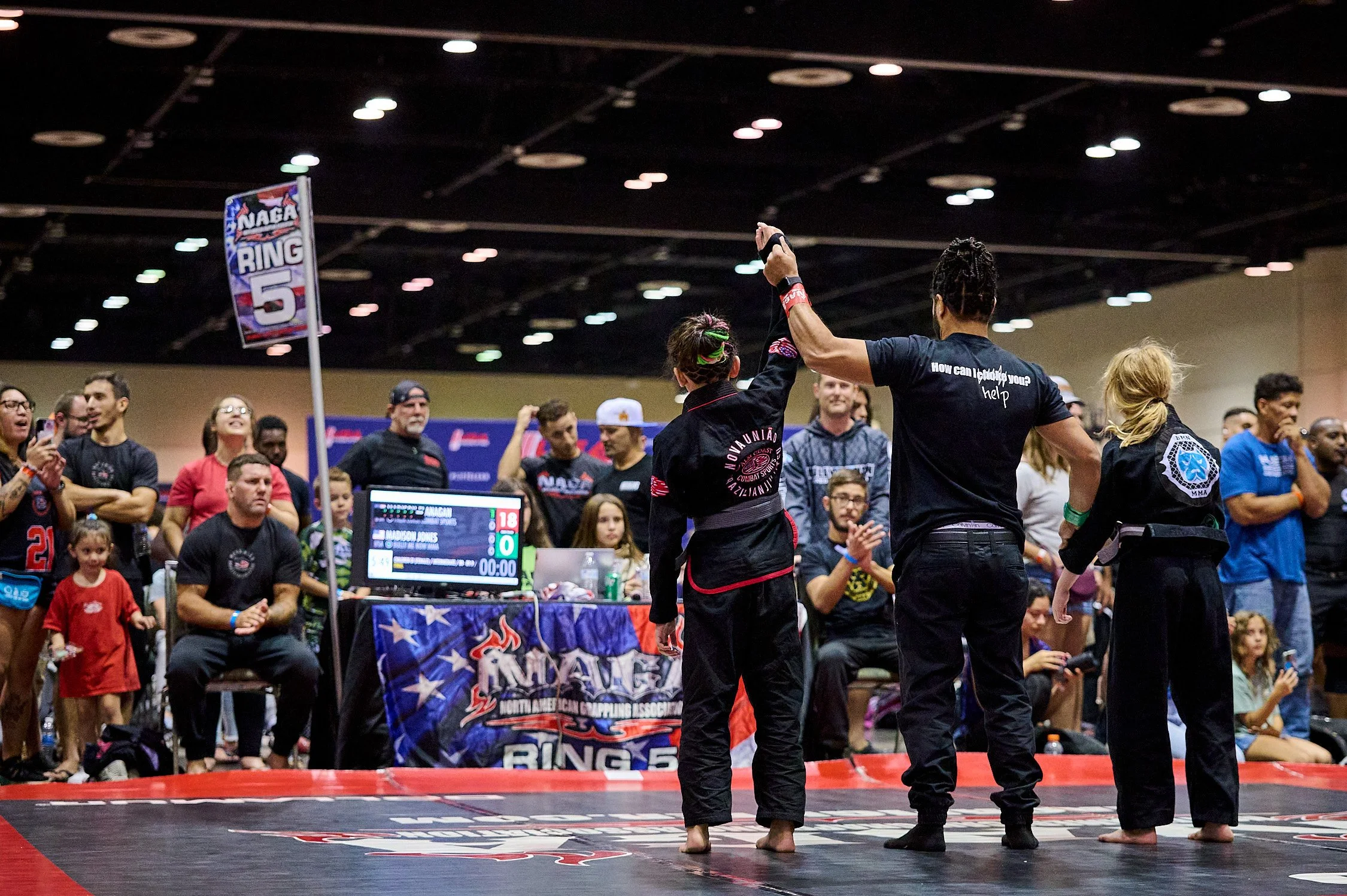 A female Brazilian Jiu-Jitsu competitor in black gi with red trim raises her arm in victory as a female referee in black t-shirt and black pants stands next to her in a mixed martial arts tournament. An audience watches in the background.