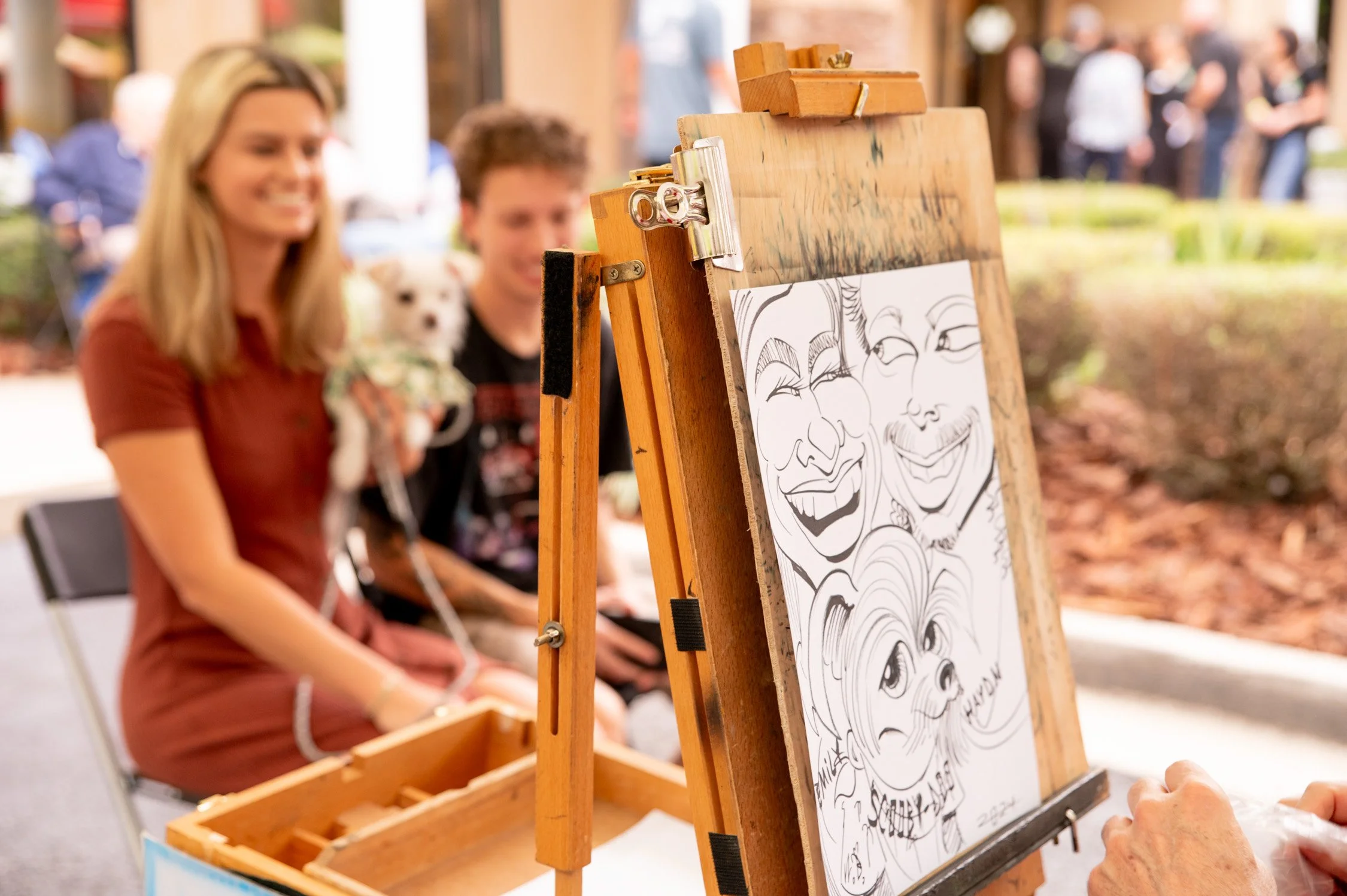 A caricature artist draws funny portraits at an outdoor event, with a smiling woman, a man, and a small dog sitting nearby. The caricatures are visible on the easel.