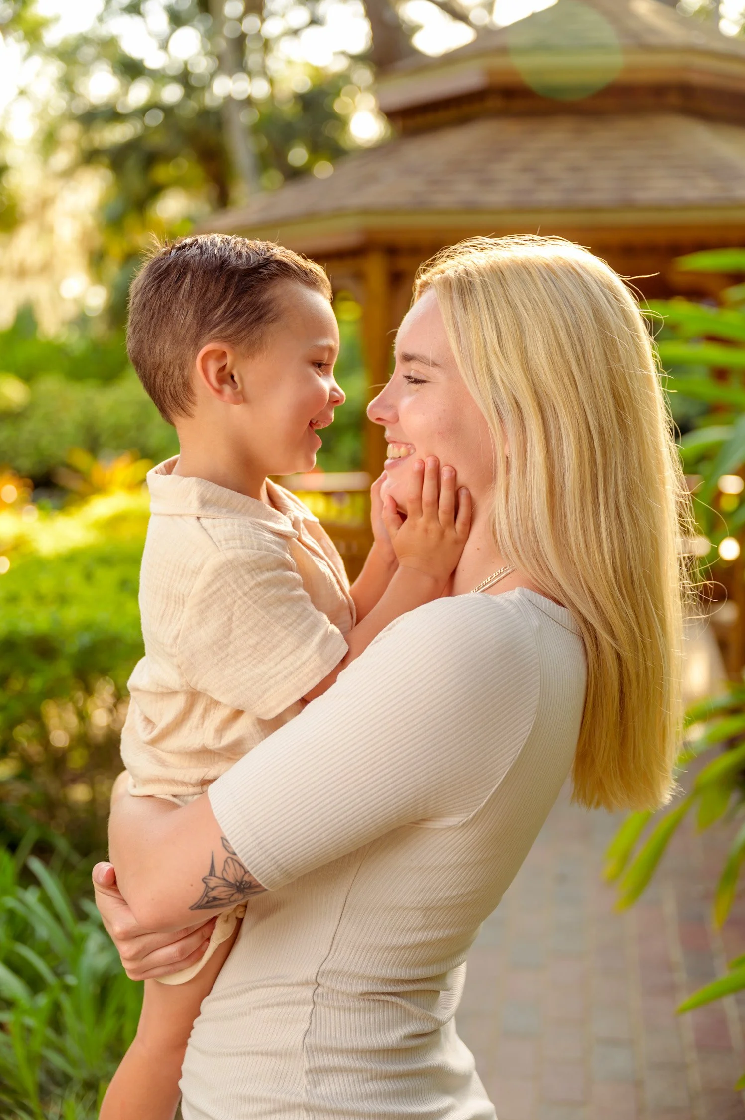 A woman with blonde hair holding a smiling young boy with short brown hair, both sharing a joyful moment outdoors.