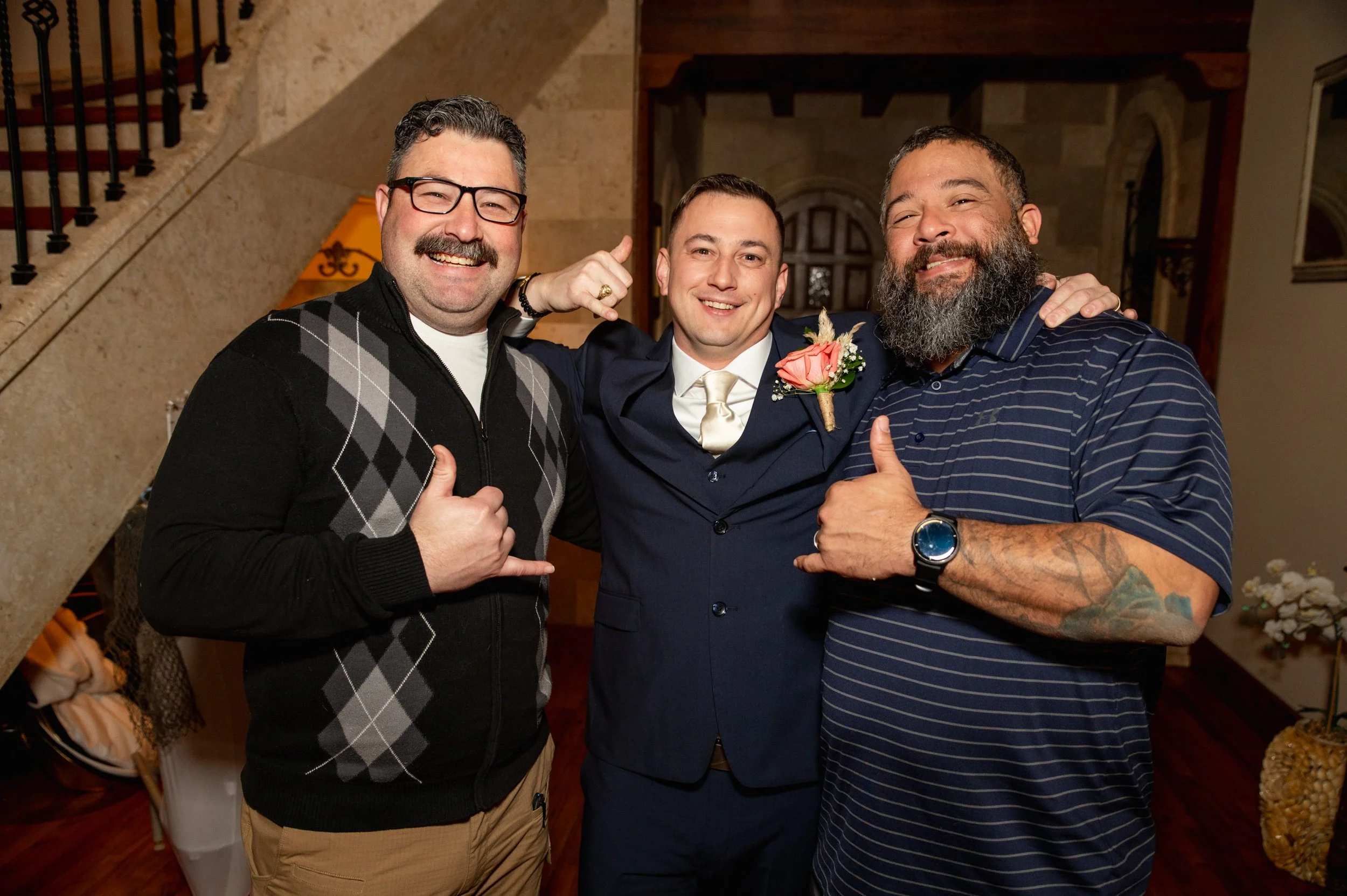 Three men joyfully posing at a wedding reception indoors, one in a black argyle sweater, one in a dark suit with a tie and boutonniere, and another in a striped shirt with tattoos.