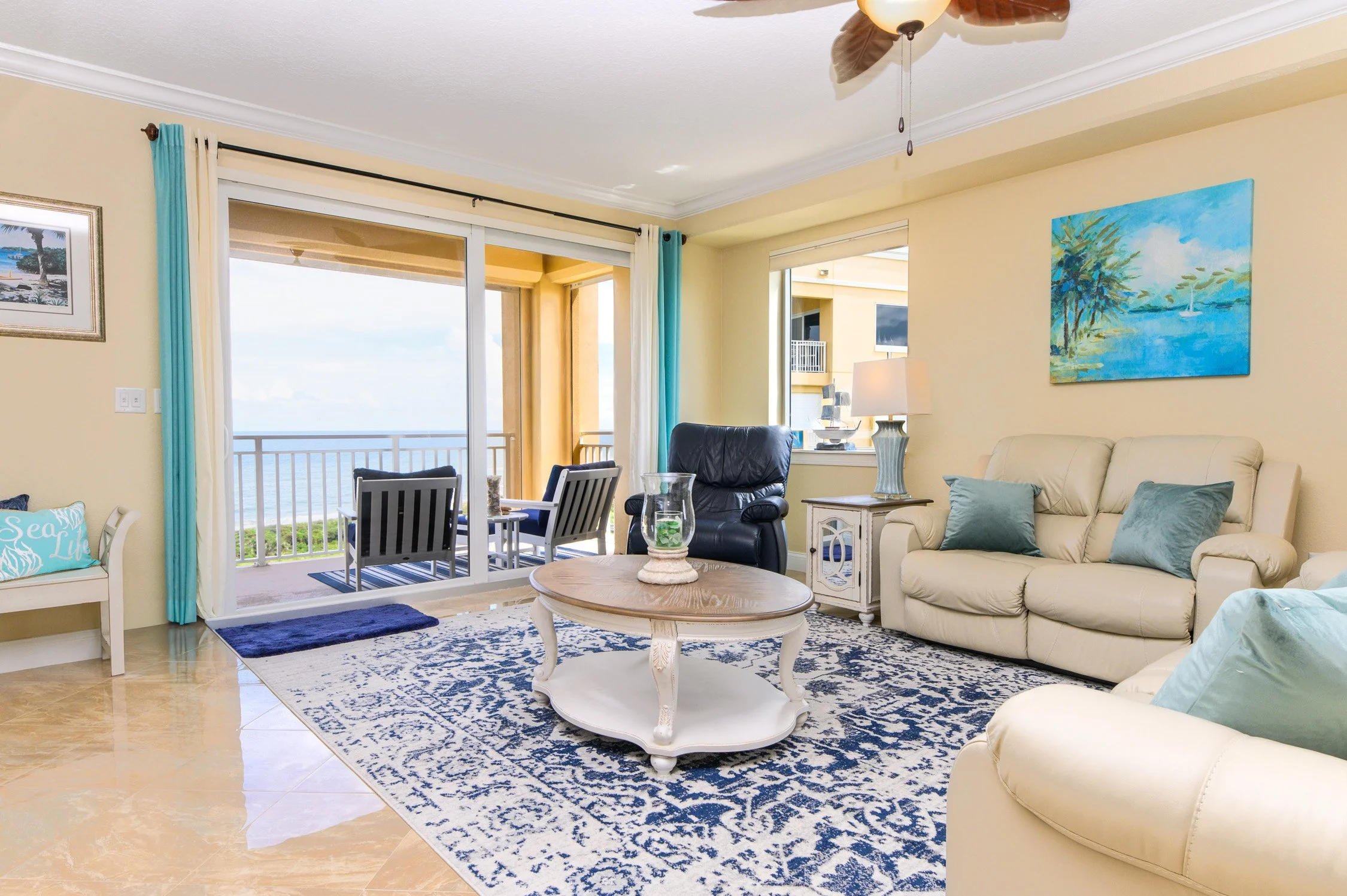Living room with beige walls, sliding glass door leading to a balcony with ocean view, blue and white patterned rug, cream leather sofa, black recliner, wooden coffee table, side table with lamp, artwork of a beach scene on the wall, and additional s