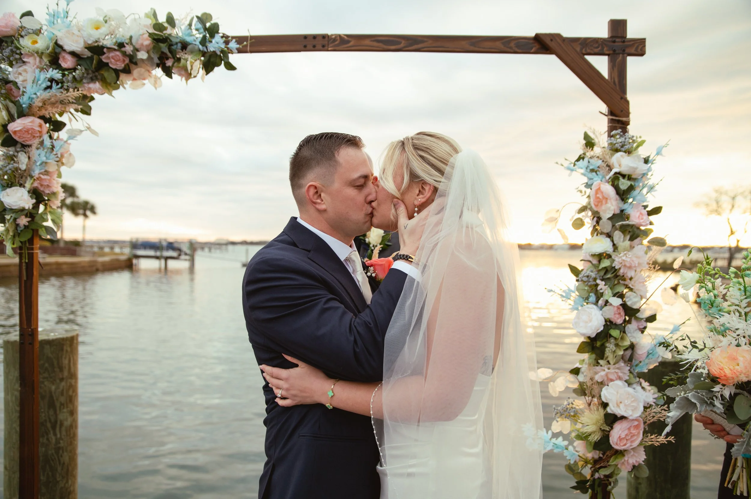 A bride and groom share a kiss during their wedding ceremony by a lake, with floral decorations and a wooden arch in the background.