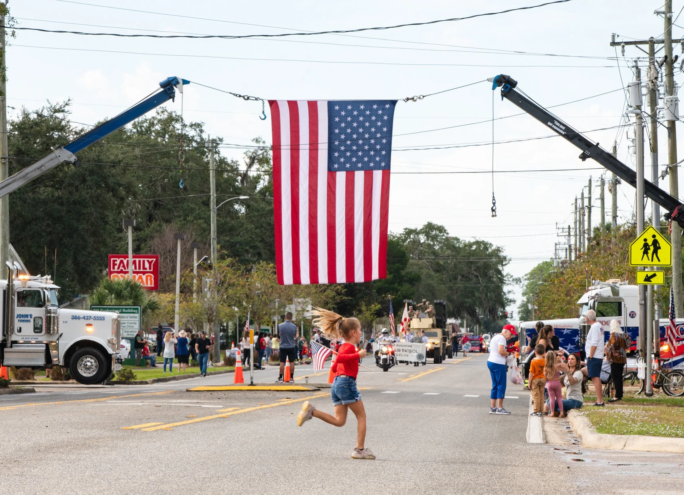 A street scene during a parade with a large American flag hanging overhead, people lining the sidewalks, a girl running with a small American flag, and parade floats and vehicles in the background.