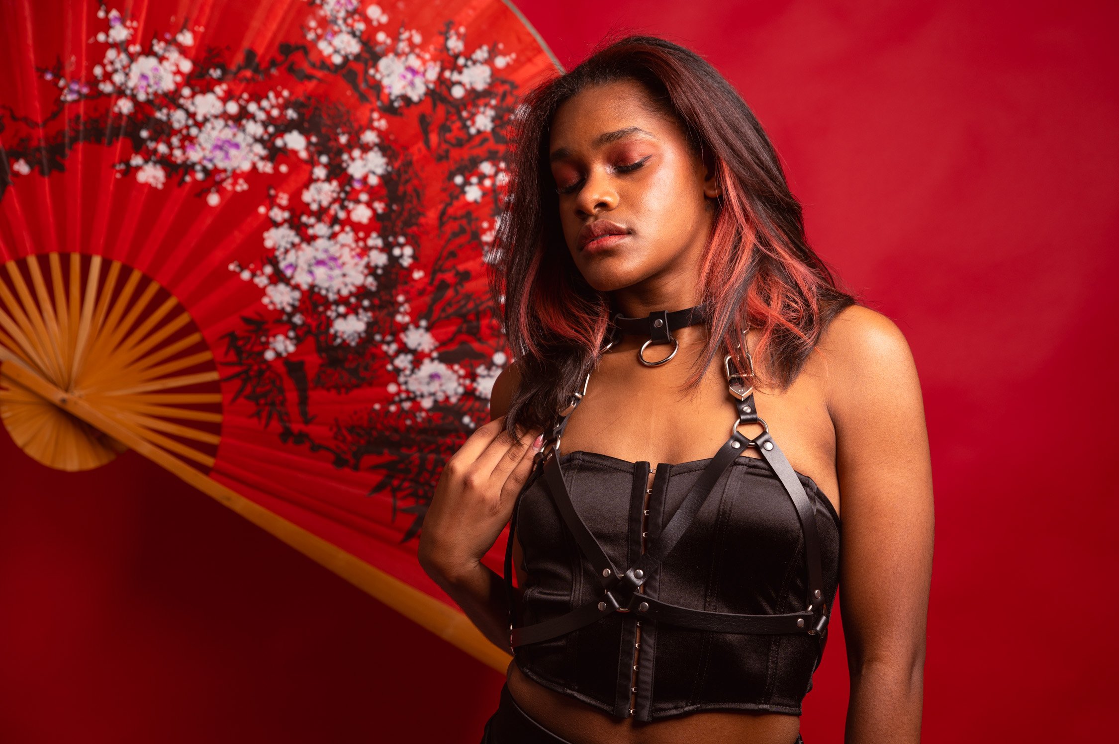 A young woman with dark hair and reddish highlights, wearing a black leather corset, harness, and choker, poses in front of a red background with a large decorative paper fan featuring cherry blossom design.