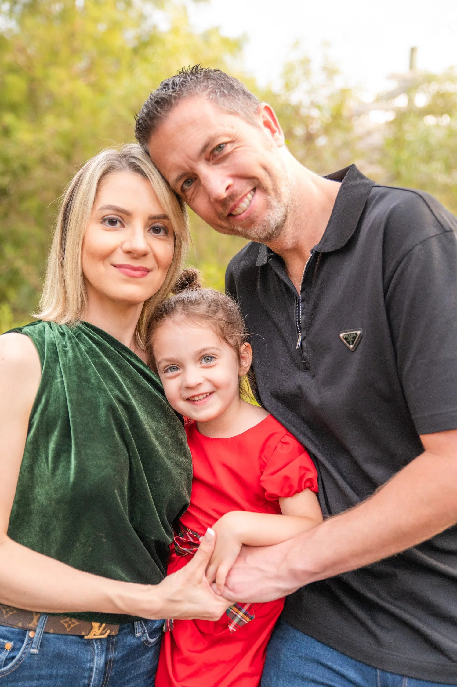 A smiling family of three outdoors with a woman, man, and young girl, closely grouped together, surrounded by green trees.