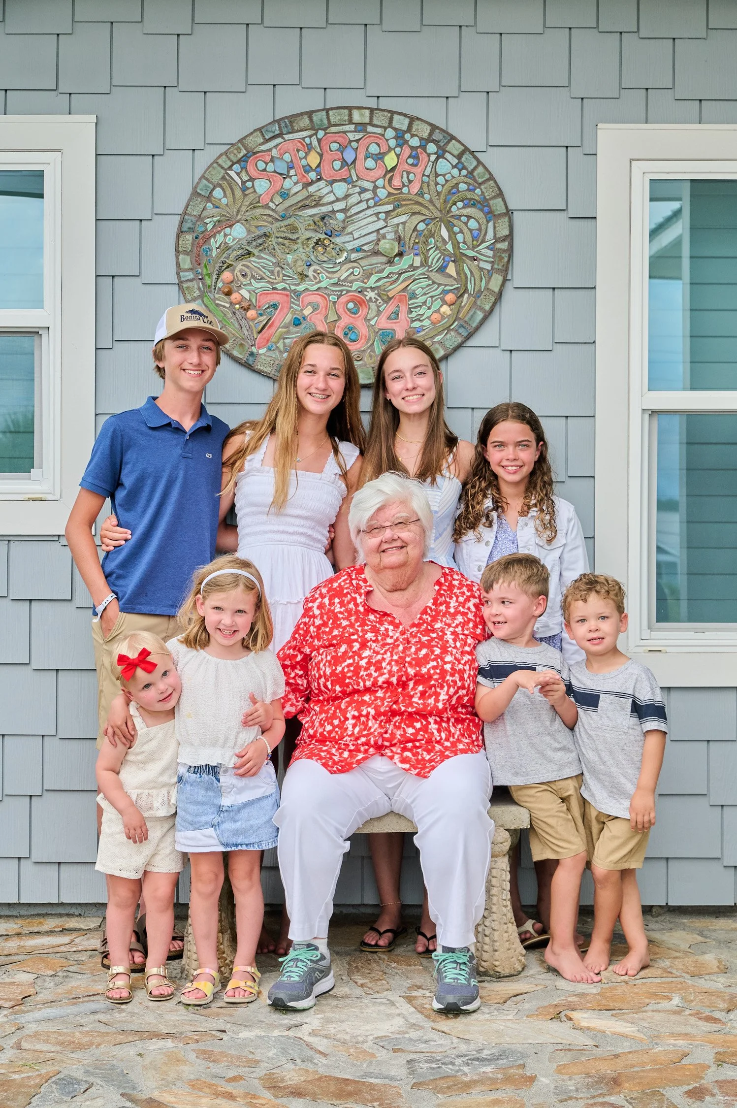 A family photo with eight children and an elderly woman, all smiling, standing outside in front of a house with grey siding and a colorful sign that reads 'STETCHI 7384' behind them.