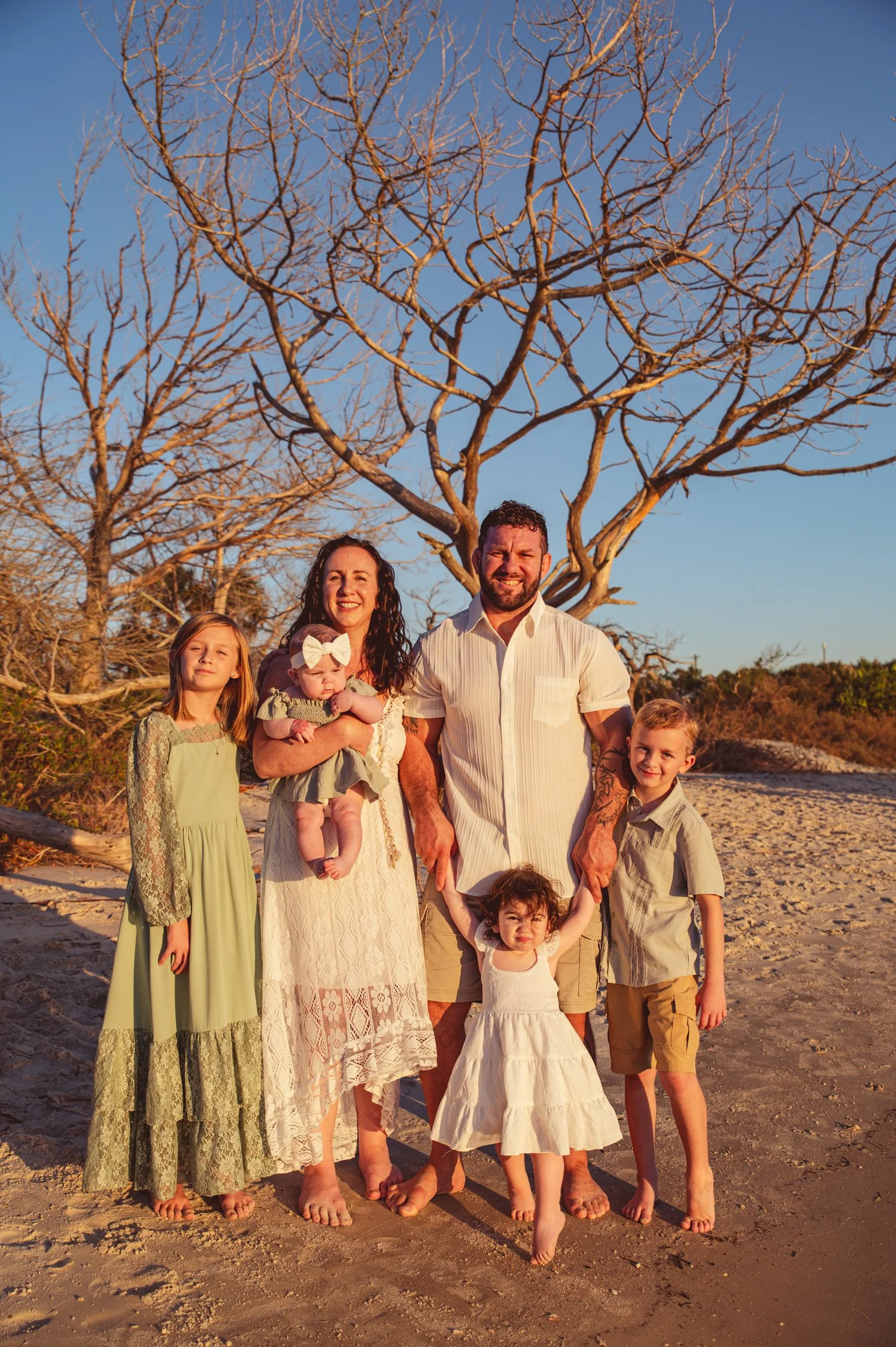 Family of six standing barefoot on sandy beach, with leafless tree and clear blue sky in the background, during sunset.