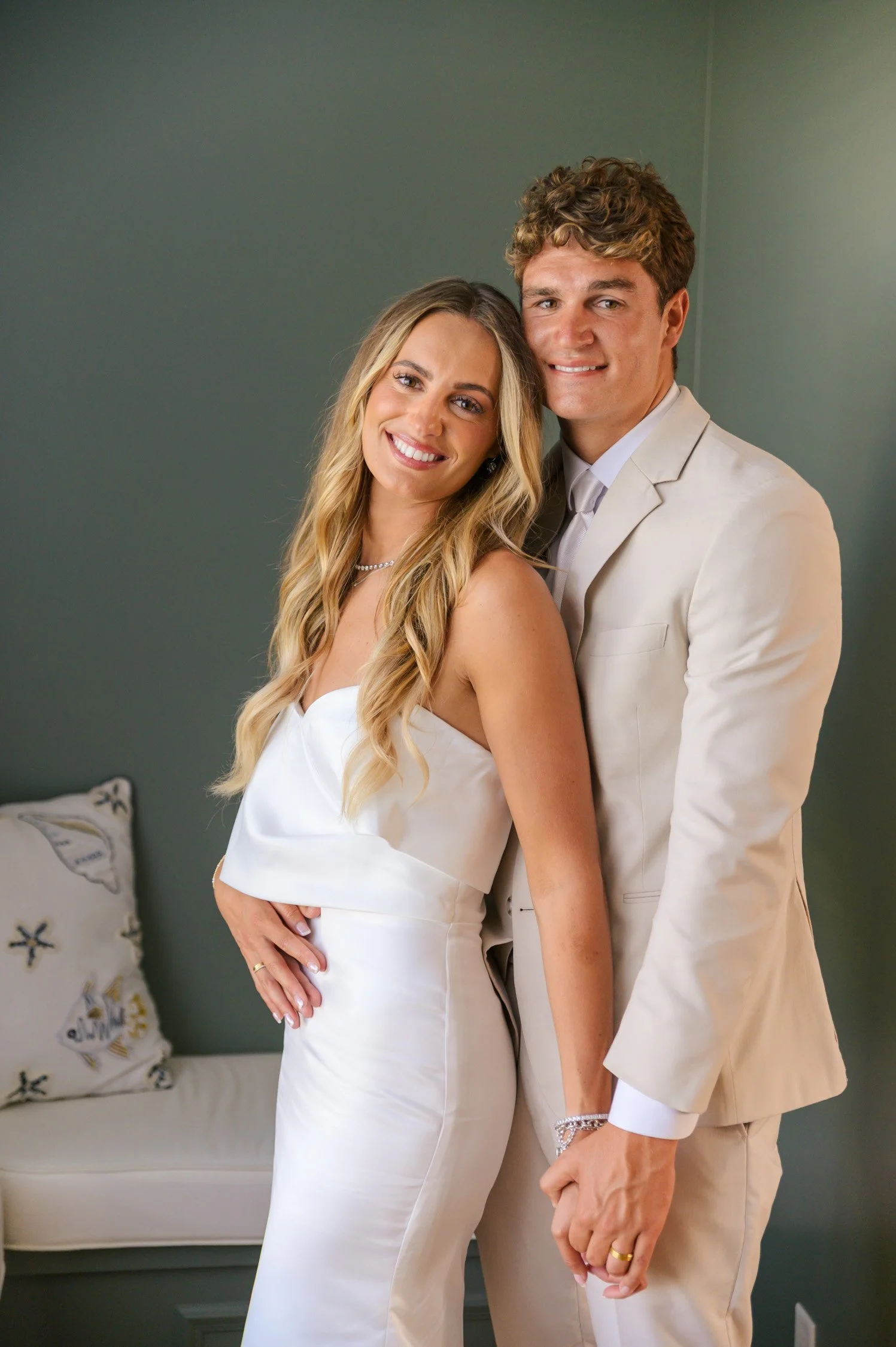 A smiling bride and groom standing close together indoors, holding hands, with a green wall and a small white sofa with a decorative pillow in the background.