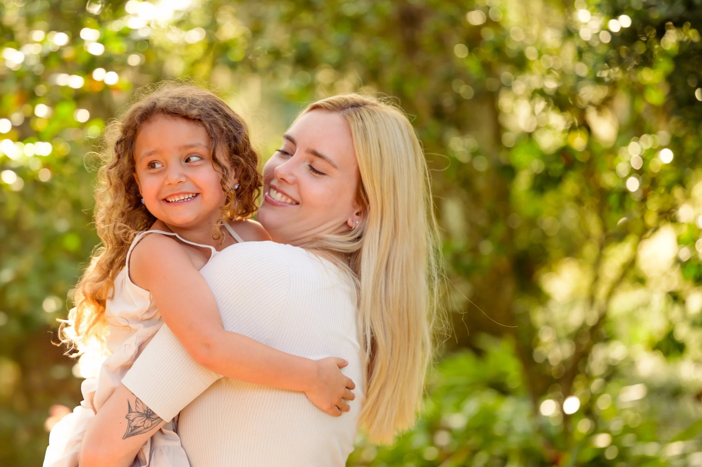 A woman with long blonde hair holding a young girl with curly brown hair outdoors during daytime. They are smiling at each other with a background of green trees and sunlight filtering through.