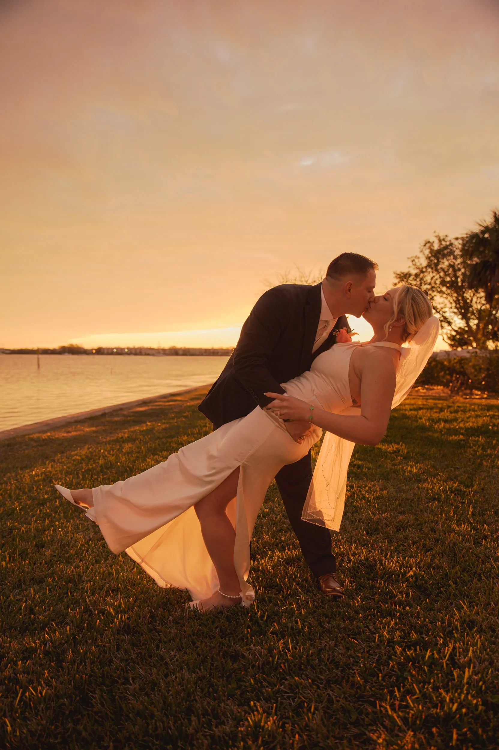 A newlywed couple kissing at sunset by the water, with the groom dipping the bride.