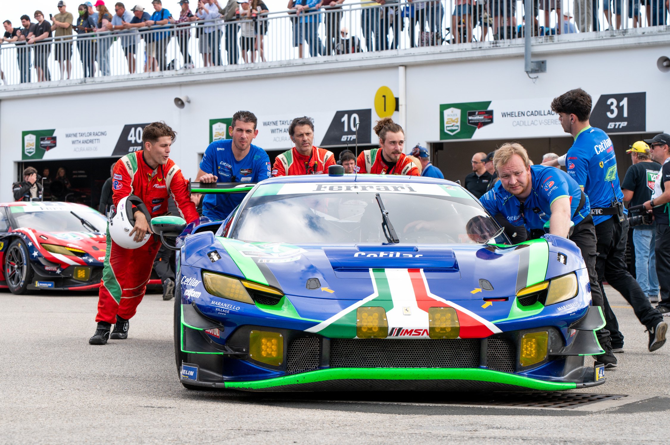 A racecar team pushing a blue, green, and white Ferrari race car in the pit lane of a racetrack, with crew members in racing suits and onlookers in the background.