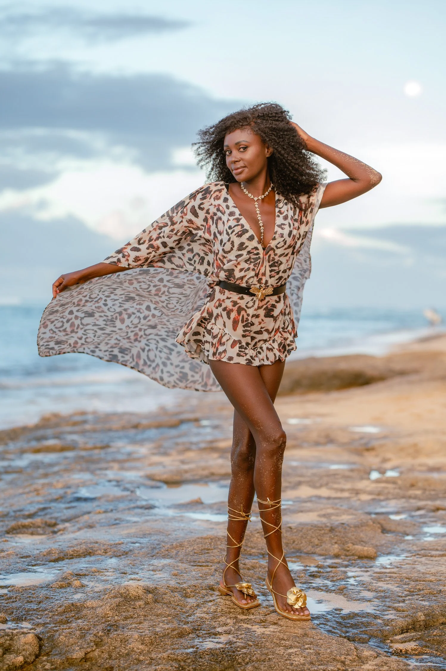 A woman in a leopard print outfit posing on a beach with the ocean in the background, wearing jewelry and sandals.