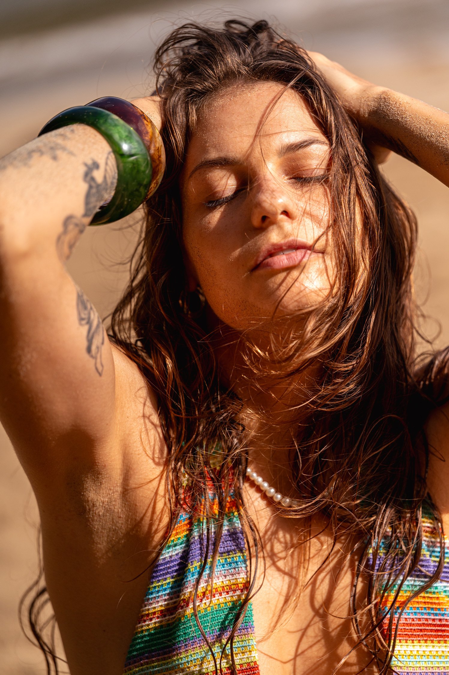 Young woman with wet hair and sand on her face and skin, relaxing on a beach with her eyes closed, wearing colorful clothing, pearl necklace, and green and brown bracelets.