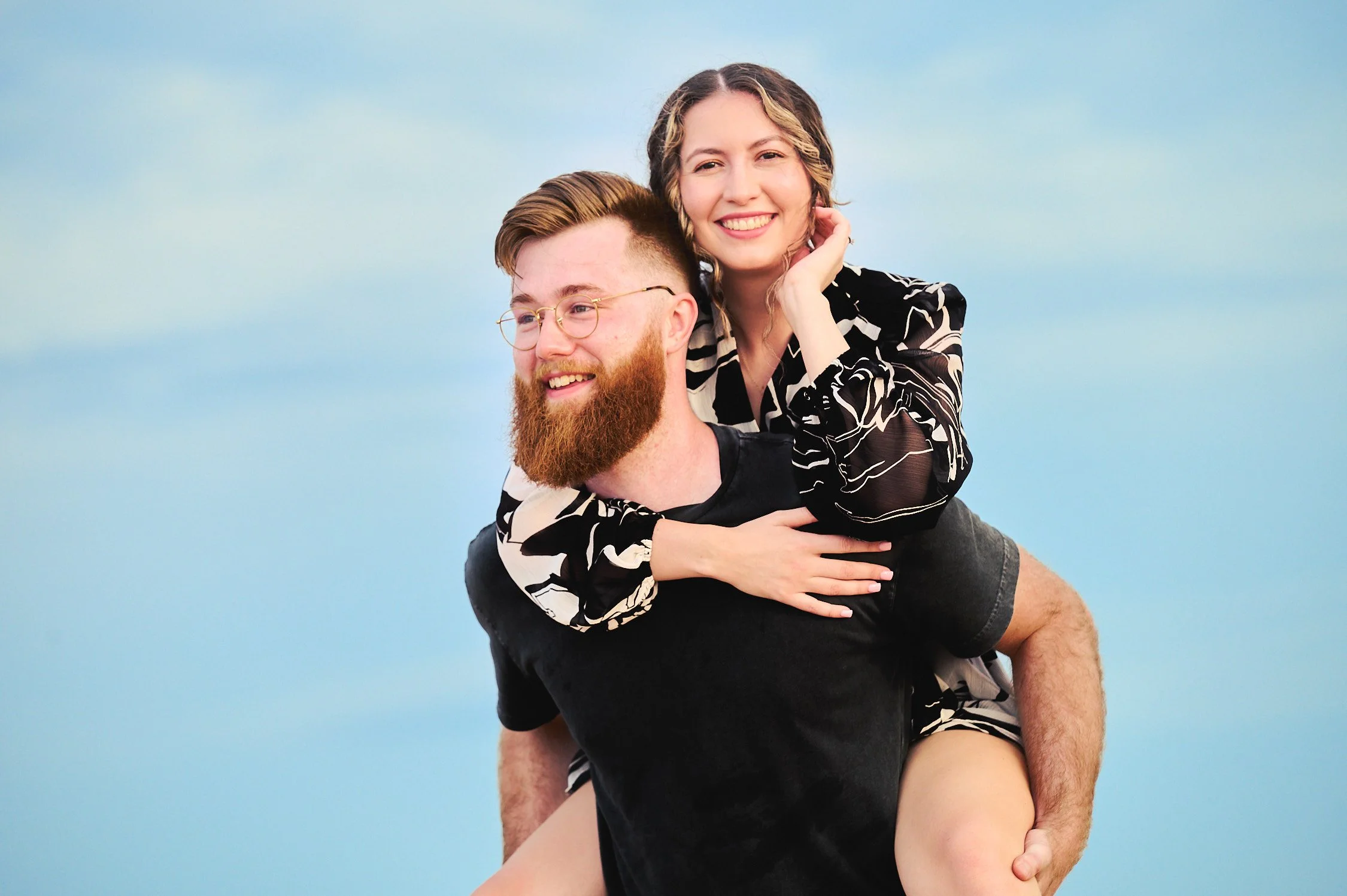 A happy couple, man with a beard and glasses, woman with curly hair, smiling, being playful outdoors with a blue sky background.