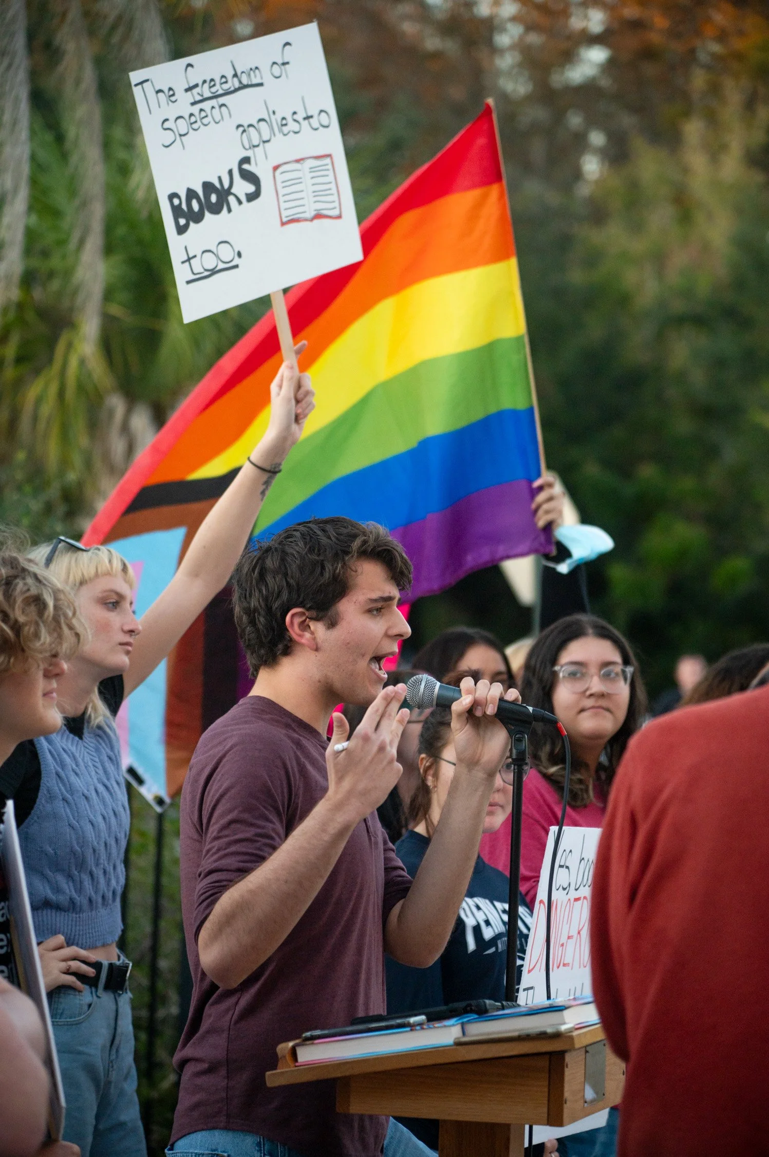 A young man speaking into a microphone at a rally, surrounded by a diverse group of people. A rainbow pride flag is visible behind him, and a sign reads, 'The freedom of speech  applied to books too.'