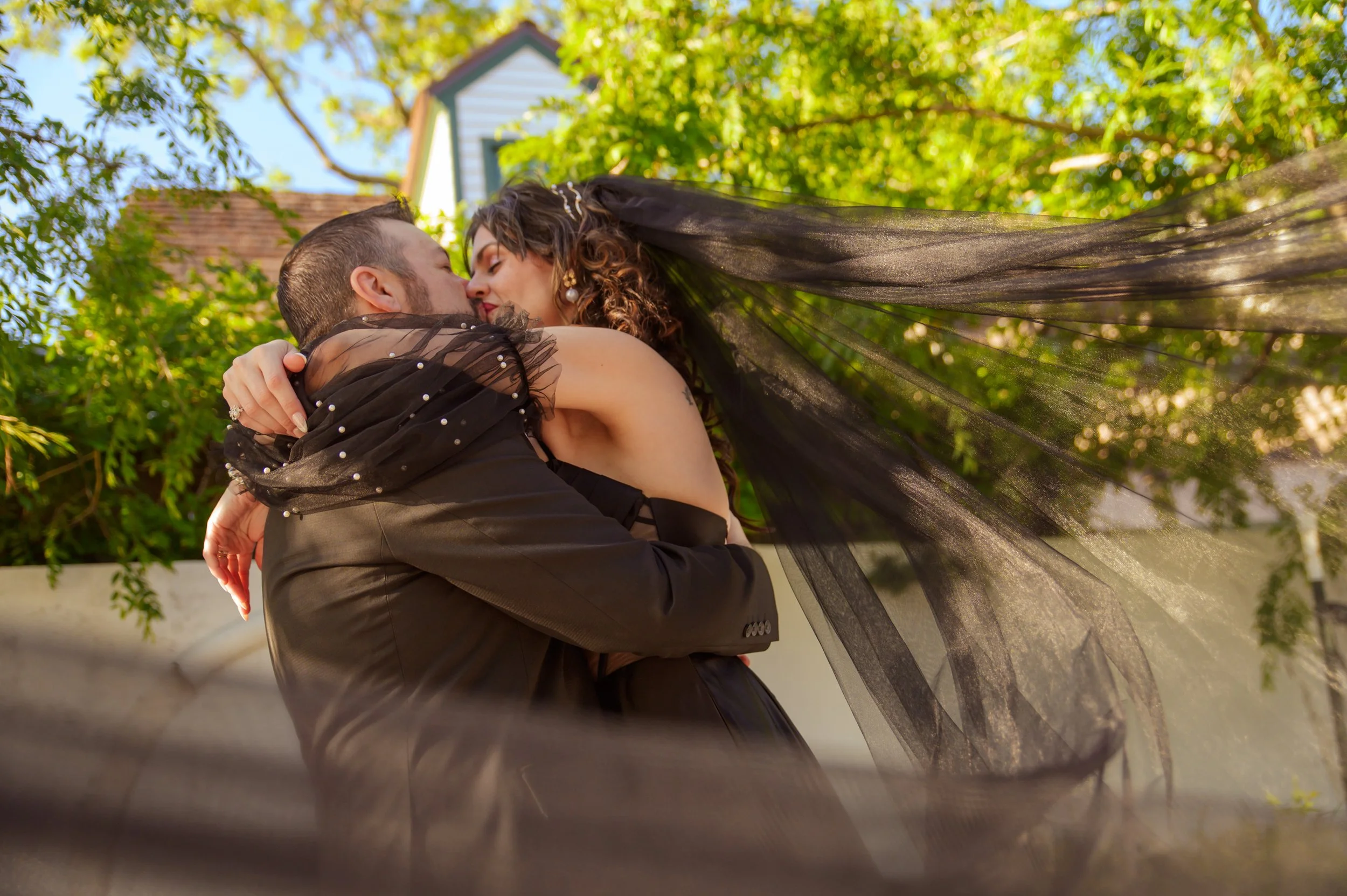 A couple embraces outdoors, with the woman wearing a black dress and veil, and the man in a black suit, surrounded by green trees and a blue sky.