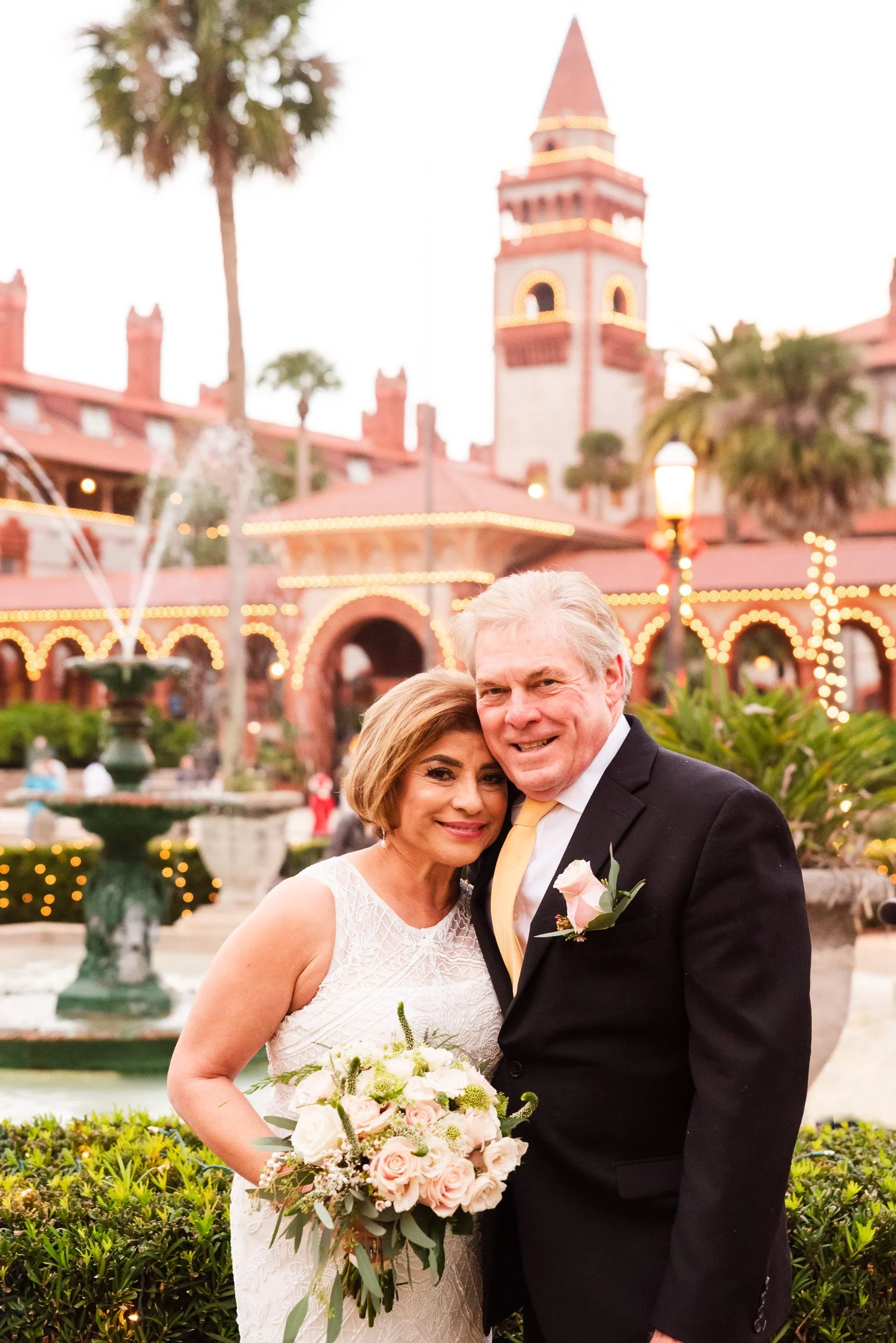 An elderly couple in formal wedding attire, standing close together, smiling, with a fountain, colorful building with lights, and palm trees in the background at sunset.