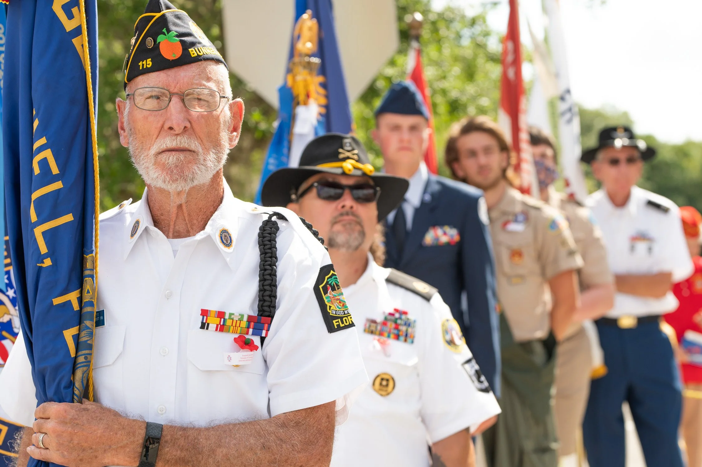A line of military veterans standing with flags during a ceremony, with the focus on an older man with a white beard and glasses holding a flag, dressed in a white uniform with medals.