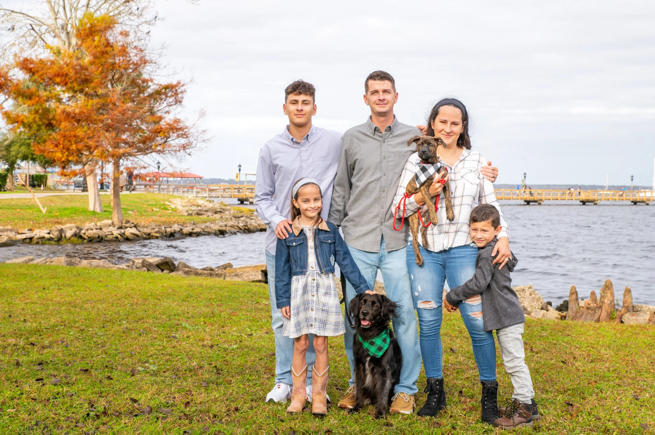 Family of six, including two adults, three children, and two dogs, standing on grass near a body of water with a bridge and trees in the background.