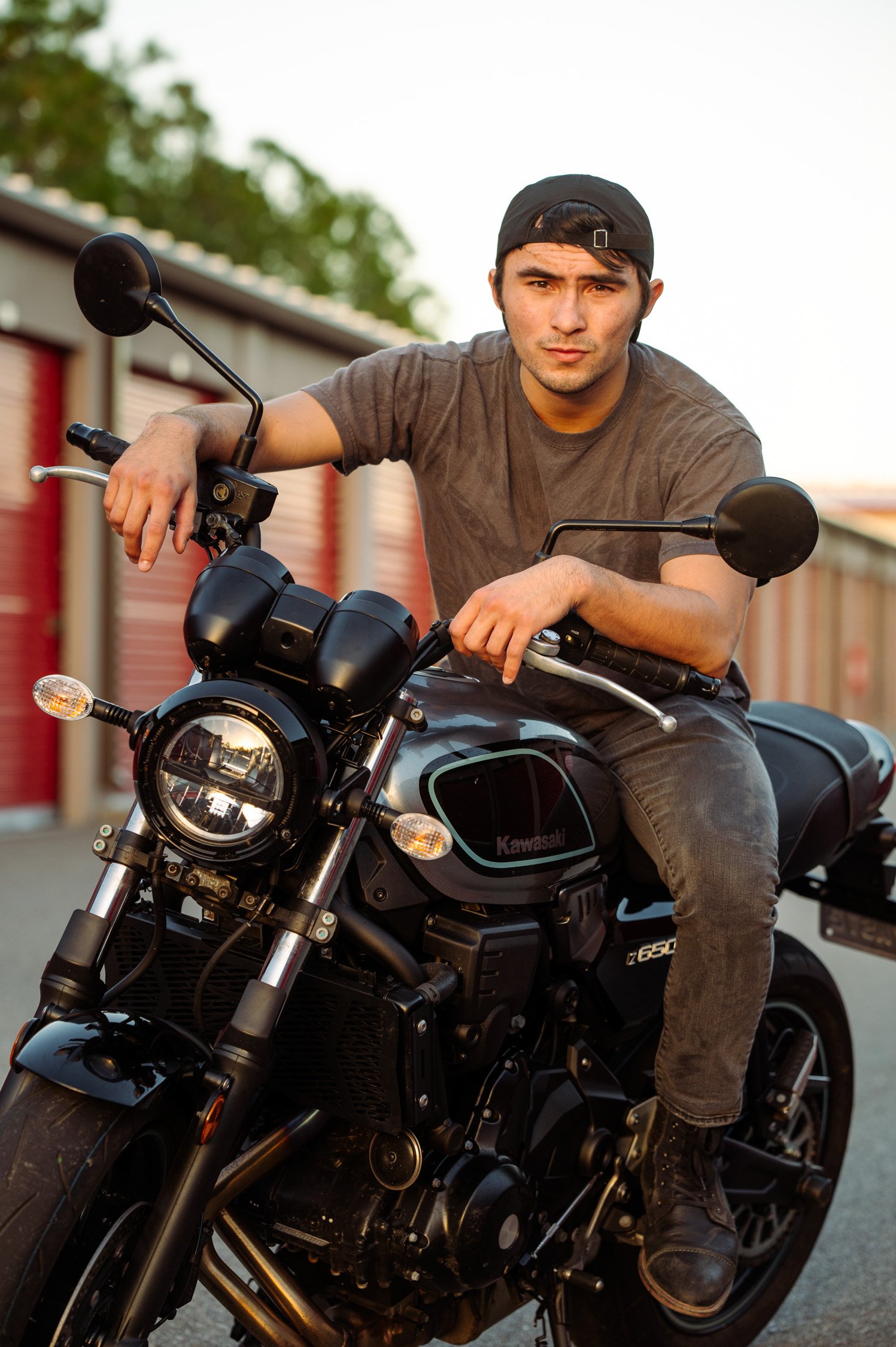 A young man in a grey t-shirt and black baseball cap sitting on a black Kawasaki motorcycle, with storage units in the background.