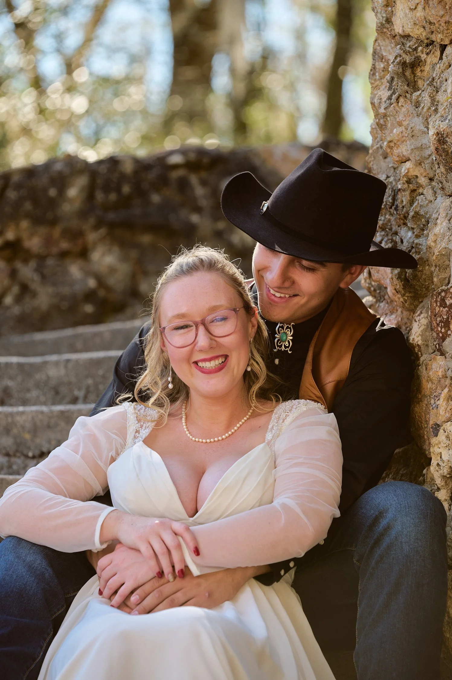 A couple dressed in Western and vintage attire sitting on stone steps outdoors, smiling and embracing each other.