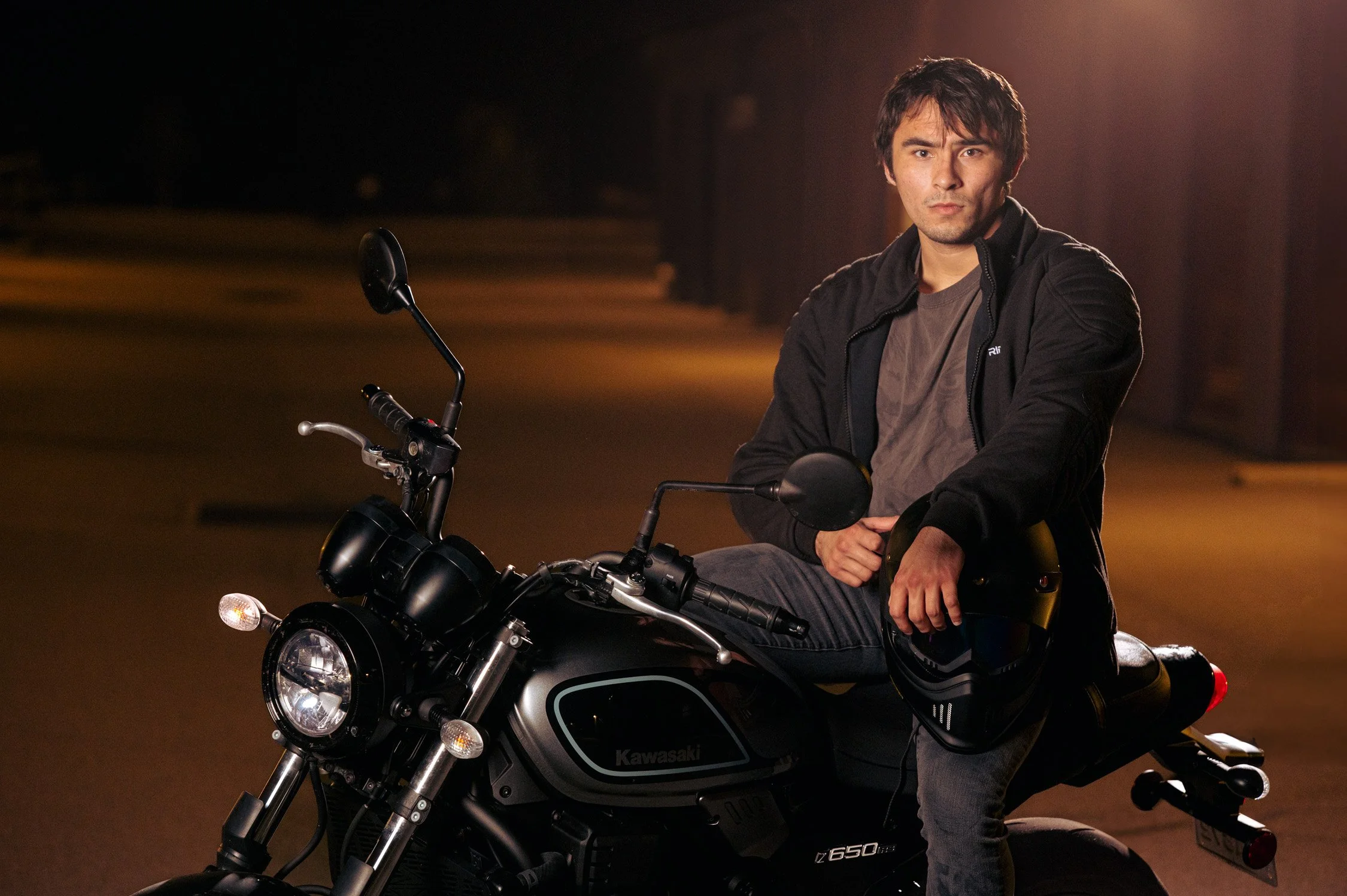A man sitting on a black Kawasaki motorcycle at night, holding a helmet in his right hand.